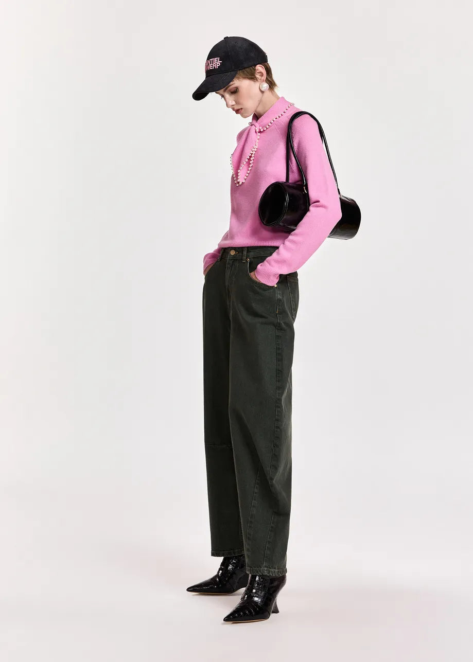 A model wearing a pink long-sleeve knit top with a ruffled collar, dark green barrel-leg jeans, black patent leather ankle boots, a black baseball cap, and a black round shoulder bag, standing against a light gray studio background.