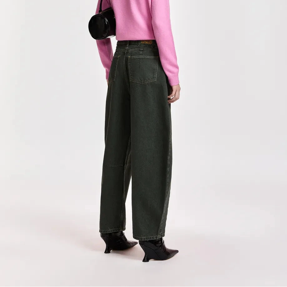 A model is shown from the back wearing a pink ribbed knit mock-neck sweater, dark olive green wide-leg jeans, black pointed-toe ankle boots, a black baseball cap, and carrying a black shoulder bag against a light gray studio background.
