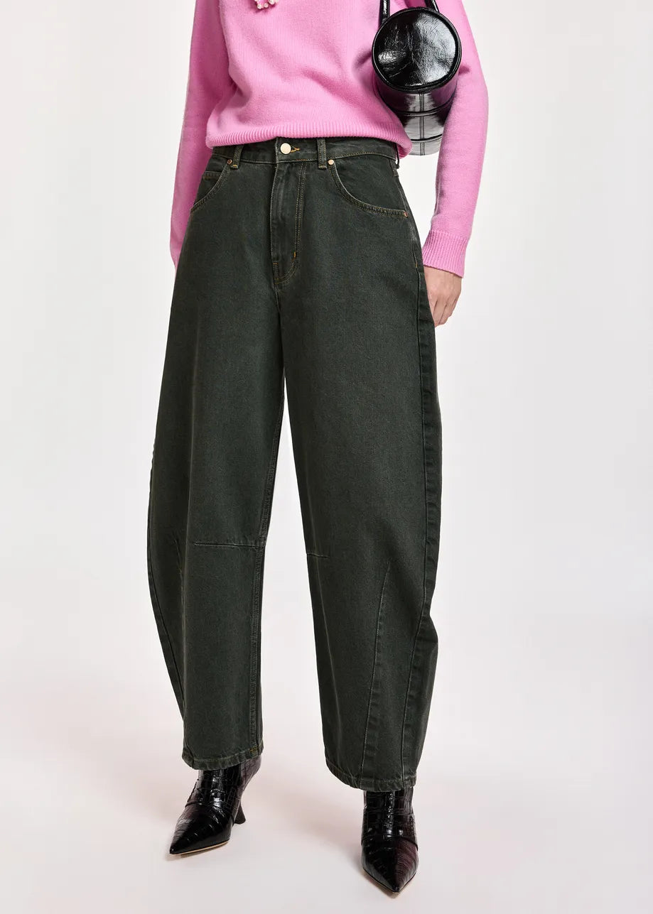 A model wearing dark olive green barrel-leg denim jeans, styled with a pink sweater, black pointed-toe boots, and a black crossbody bag against a light gray studio background.