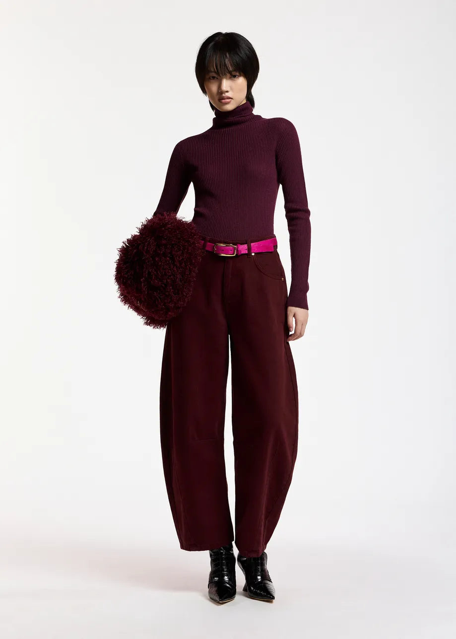 Wearing Essentiel Antwerp’s Kokos Colour Denim Barrel Leg Jeans in burgundy, styled with a matching turtleneck, black boots, a dark red fluffy bag, and a magenta belt, this person stands against a plain white background.