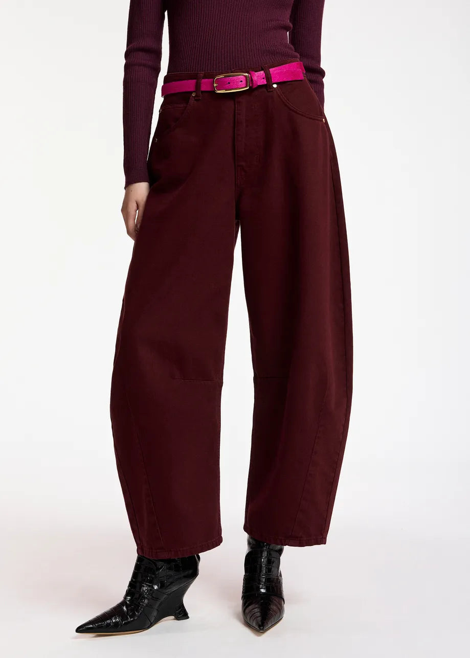A person stands against a white background wearing the Essentiel Antwerp Kokos Colour Denim Barrel Leg Jeans in burgundy, styled with a matching burgundy top, a bright pink belt, and pointed black boots.