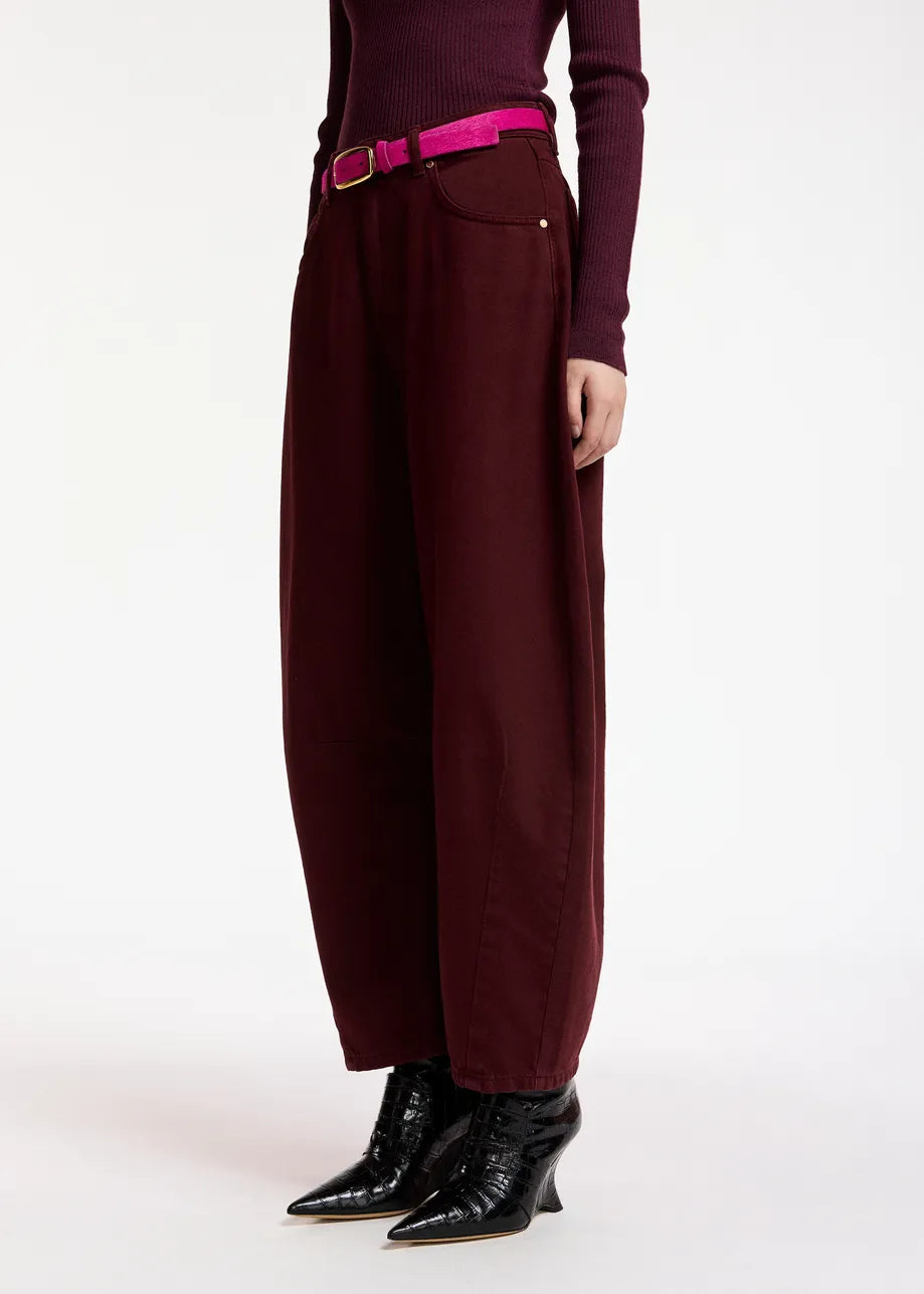 A person wearing Essentiel Antwerp’s Kokos Colour Denim Barrel Leg Jeans in burgundy, paired with a matching long-sleeve top, a bright pink belt, and black pointed boots, stands against a plain white background.