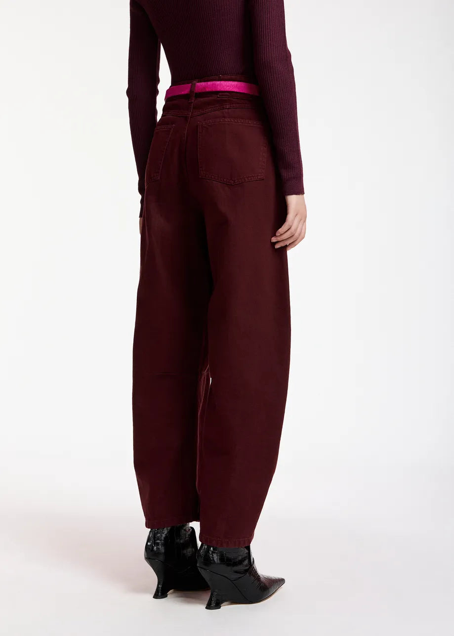 A person is seen from the back against a plain white background, wearing the Essentiel Antwerp Kokos Colour Denim Barrel Leg Jeans in Burgundy—a maroon, high-rise barrel-leg style—paired with a matching long-sleeve top made from 100% organic cotton, a pink belt, and black pointed boots.