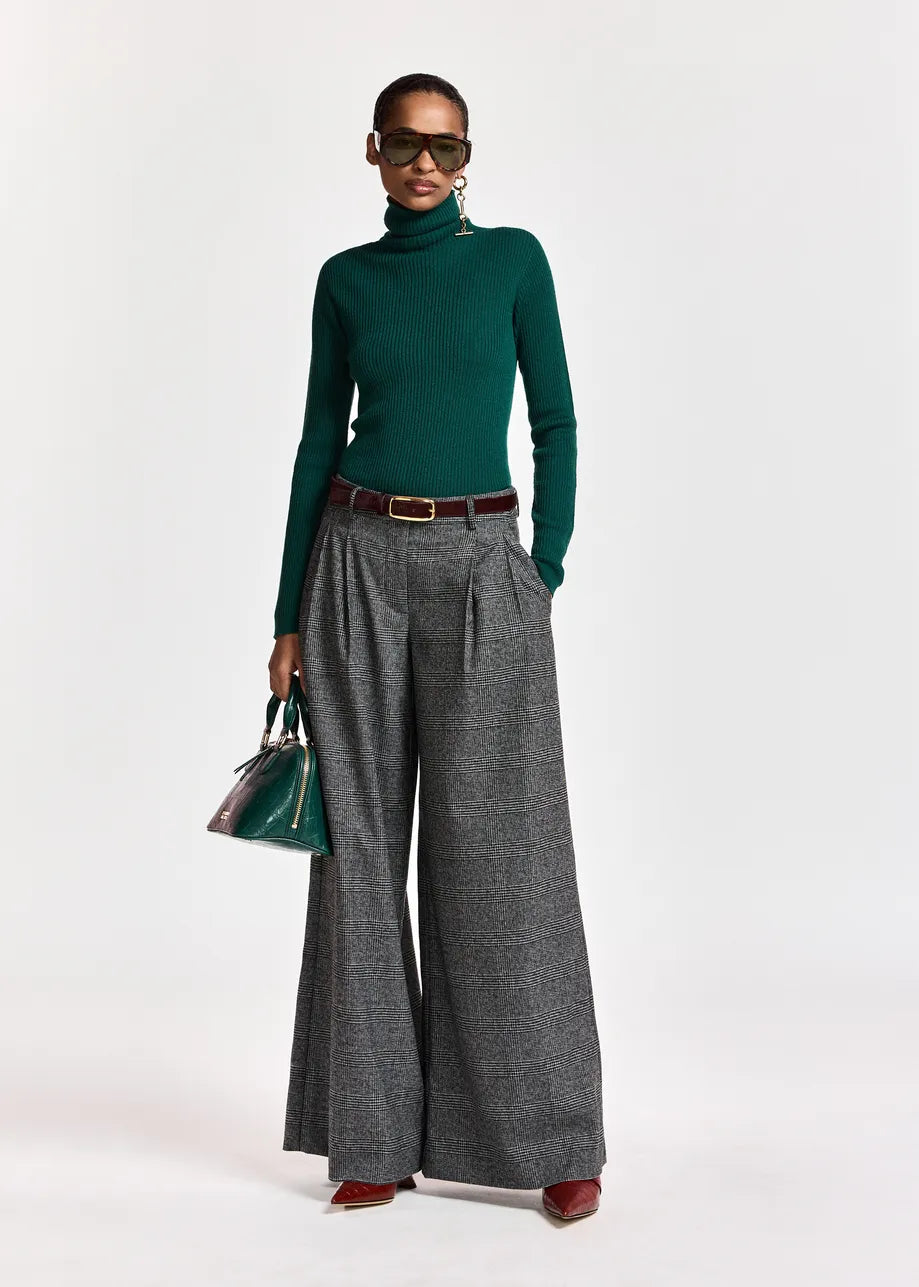 A person is wearing large sunglasses, a green turtleneck, and the Essentiel Antwerp Kollection Checkered Wide-Leg Pants in black and grey—expertly crafted from recycled polyester. Paired with brown shoes and a green handbag, they stand against a plain white background. The relaxed silhouette of the trousers exemplifies effortless style with an eco-conscious appeal.