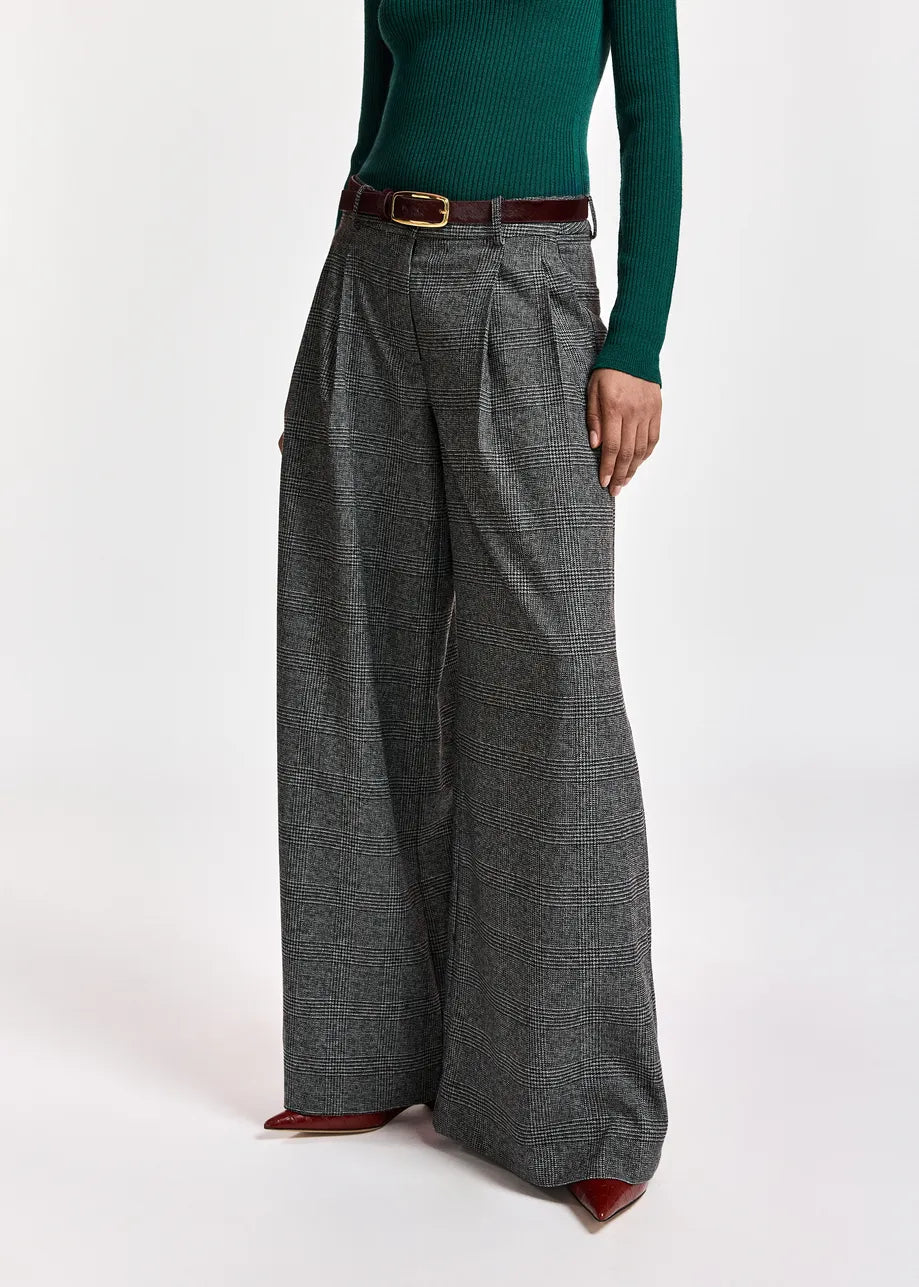 A person wearing the Essentiel Antwerp Kollection Checkered Wide-Leg Pants in Black/Grey, crafted from recycled polyester, paired with a green ribbed sweater, a brown belt, and brown pointed shoes stands against a plain white background. The pants’ relaxed silhouette adds effortless style to the overall look.