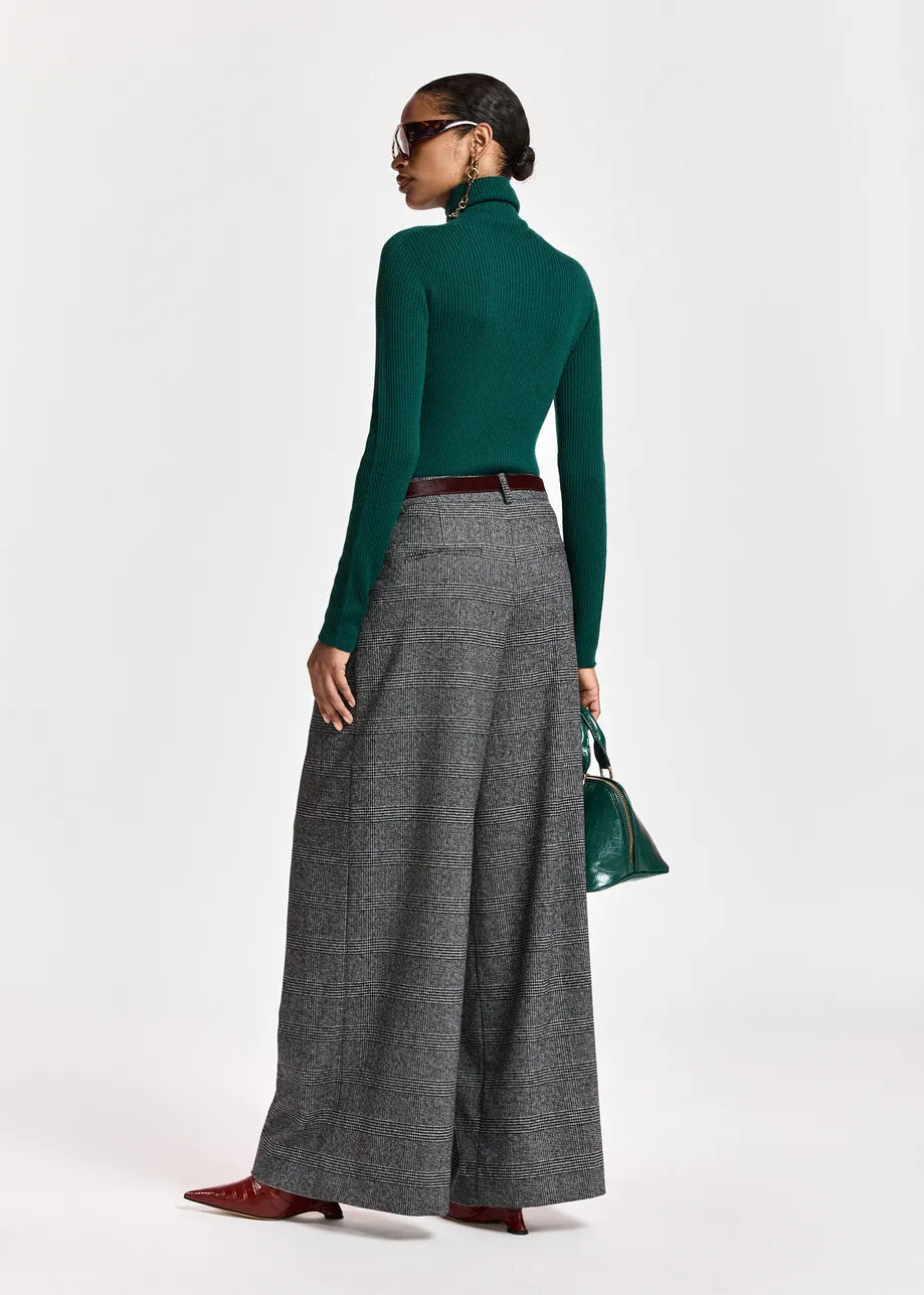 A person stands facing away, wearing a green turtleneck sweater and Essentiel Antwerp Kollection Checkered Wide-Leg Pants in black/grey made from recycled polyester, paired with red shoes, sunglasses, and holding a green handbag. The outfit showcases a relaxed silhouette with effortless style.
