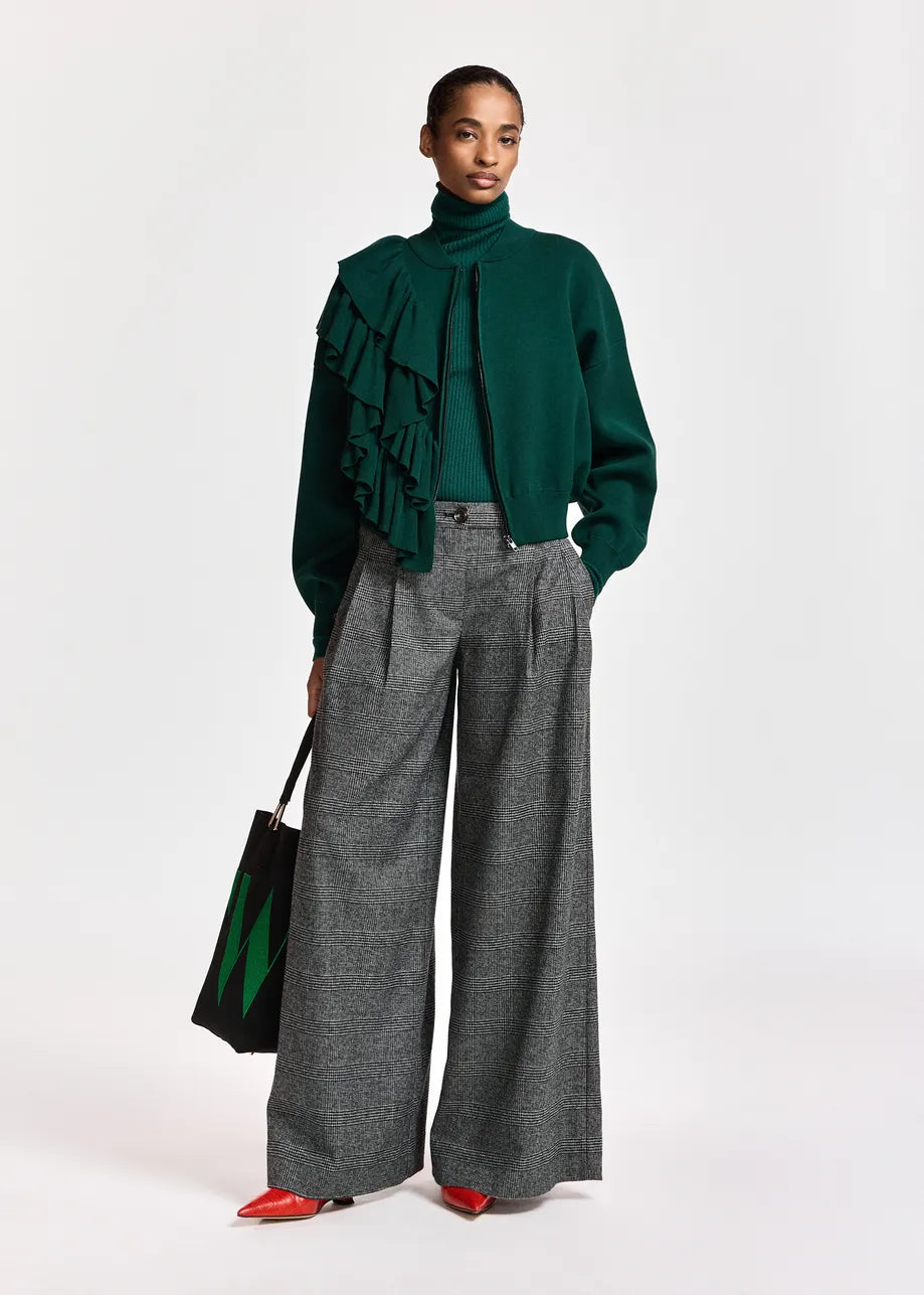Dressed in a green turtleneck with ruffle detail, she pairs the Essentiel Antwerp Kollection Checkered Wide-Leg Pants in black and grey with red pointed shoes and holds a black tote bag featuring a green “W” motif. The relaxed silhouette of her trousers completes her effortlessly chic look against the plain backdrop.