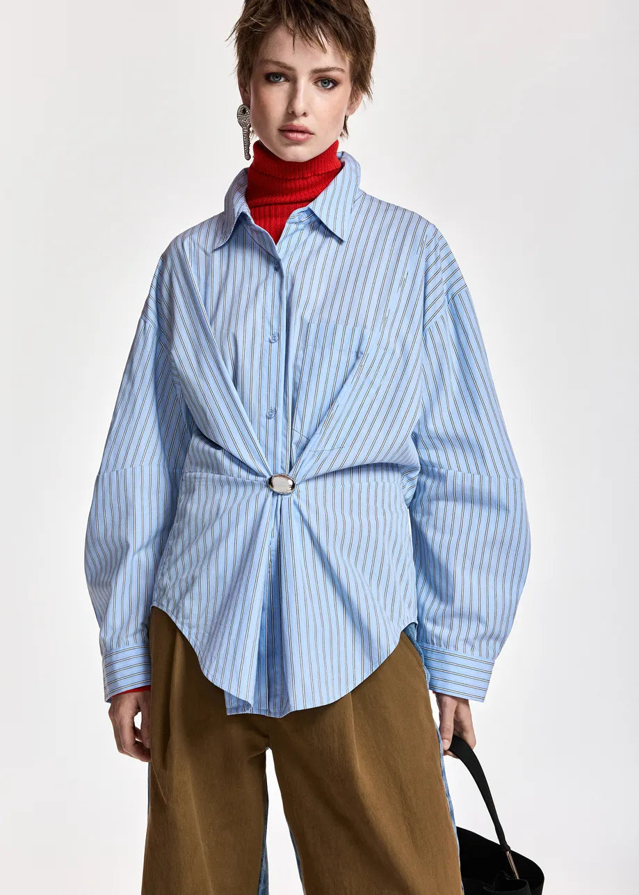 A model wearing a light blue and white vertically striped button-down shirt styled with a red turtleneck, brown wide-leg trousers, and a black handbag against a plain light gray background.
