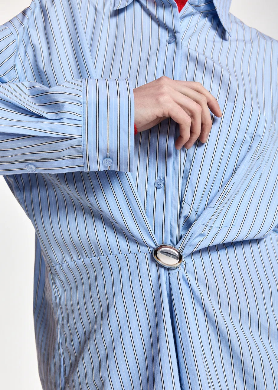 Close-up of a person wearing a light blue long-sleeve button-up shirt with thin vertical dark stripes and a large round metal button at the waist, with one hand adjusting the front.