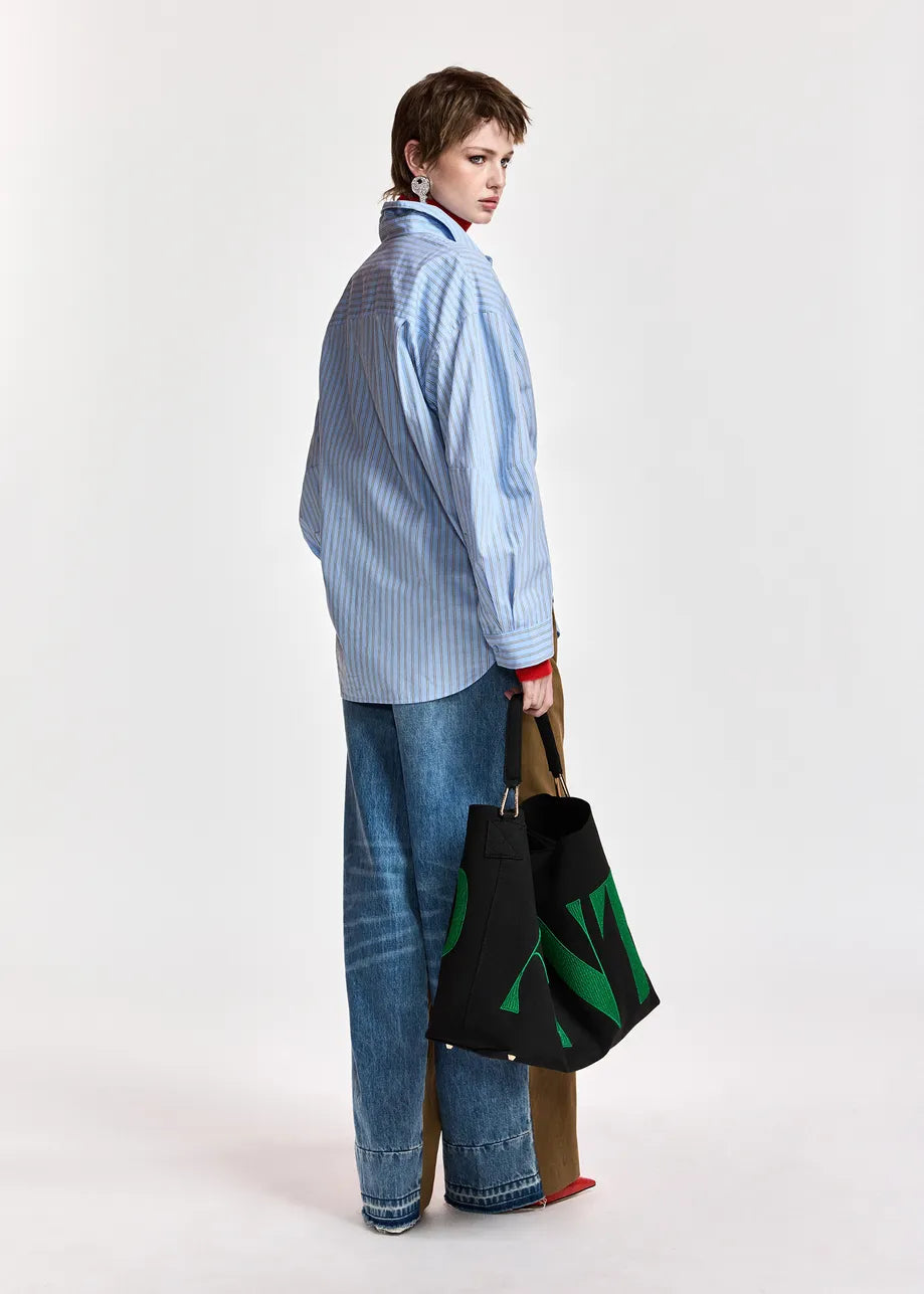 A model is shown from the back wearing a light blue and white striped button-down shirt, wide-leg blue jeans, and red shoes, while holding a black canvas tote bag with green abstract patterns and brown straps against a plain white studio background.