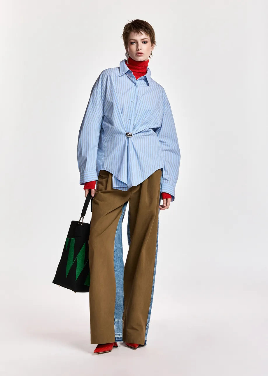 A model stands against a light gray studio background wearing a light blue and white vertically striped button-up shirt tied at the waist, wide-leg brown pants with light blue side panels, a red turtleneck, and red pointed shoes, while holding a black tote bag with a green graphic.