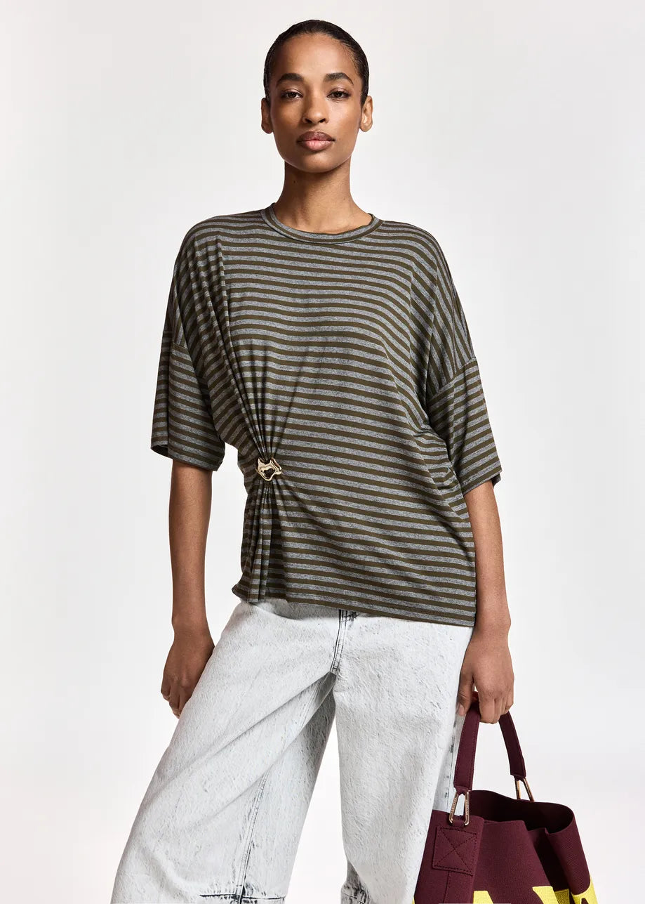 A person stands against a plain background wearing the Essentiel Antwerp Kriss Striped T-Shirt with Brooch in khaki, styled with light-wash jeans and holding a burgundy bag.