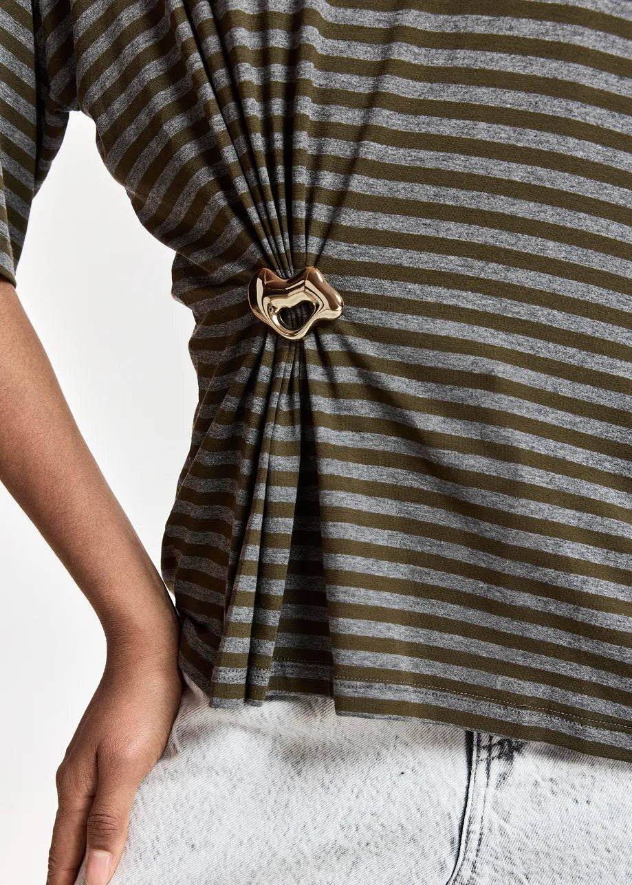 A person wears the Essentiel Antwerp Kriss Striped T-Shirt with Brooch in khaki, styled with light-colored denim jeans for a relaxed fit.