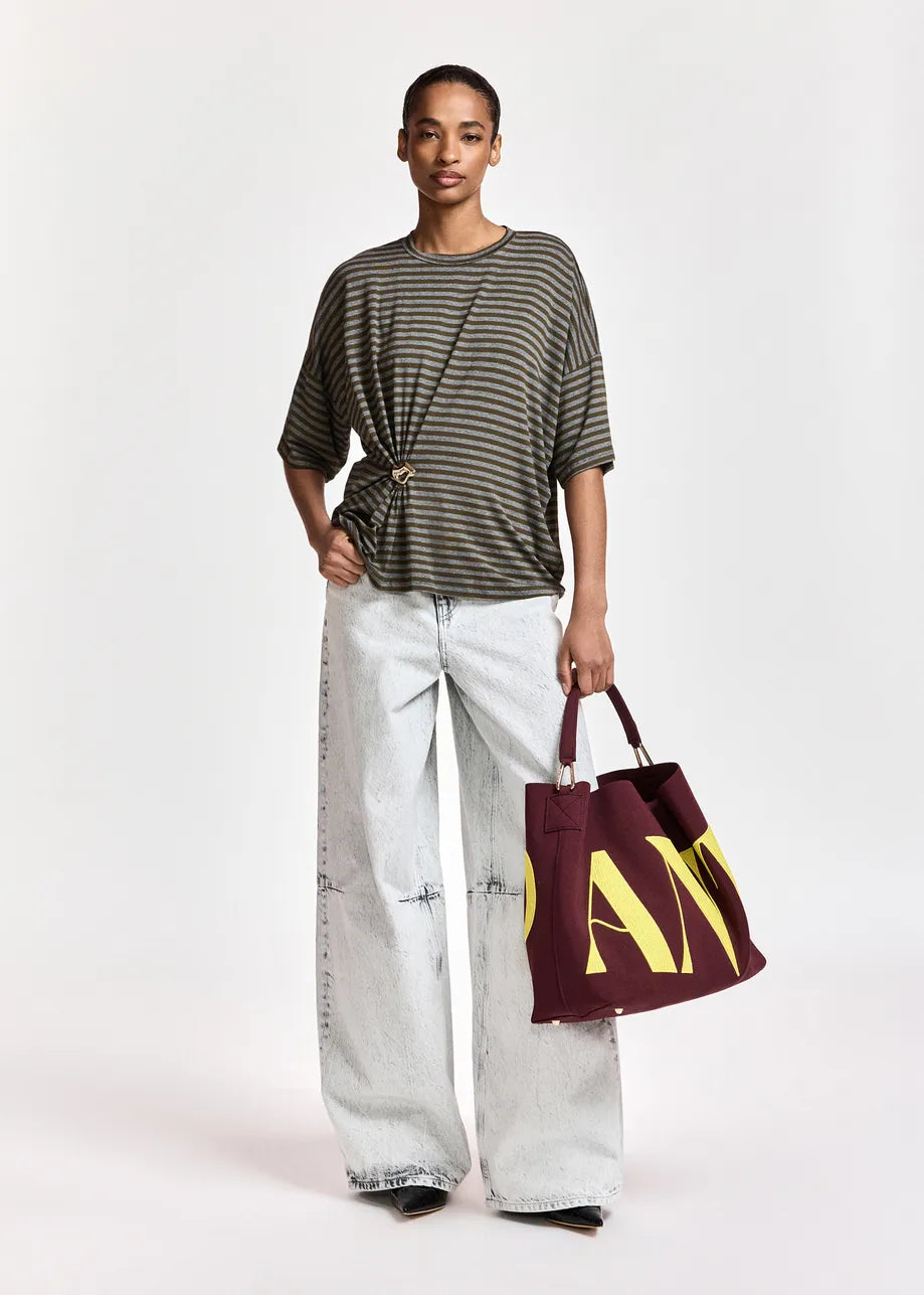 Wearing the Essentiel Antwerp Kriss Striped T-Shirt with Brooch in khaki, styled with wide-leg light-wash jeans and carrying a large maroon tote bag featuring yellow letters, this person stands against a plain light background for a relaxed fit ensemble.