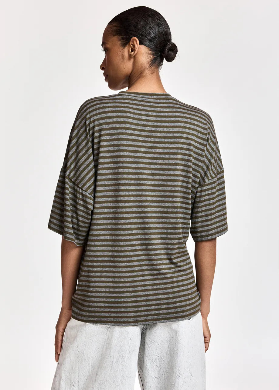 Person standing with back to the camera, wearing the Essentiel Antwerp Kriss Striped T-Shirt with Brooch in khaki—an oversized, relaxed-fit tee crafted from a soft viscose blend—paired with light-colored pants, against a plain white background.