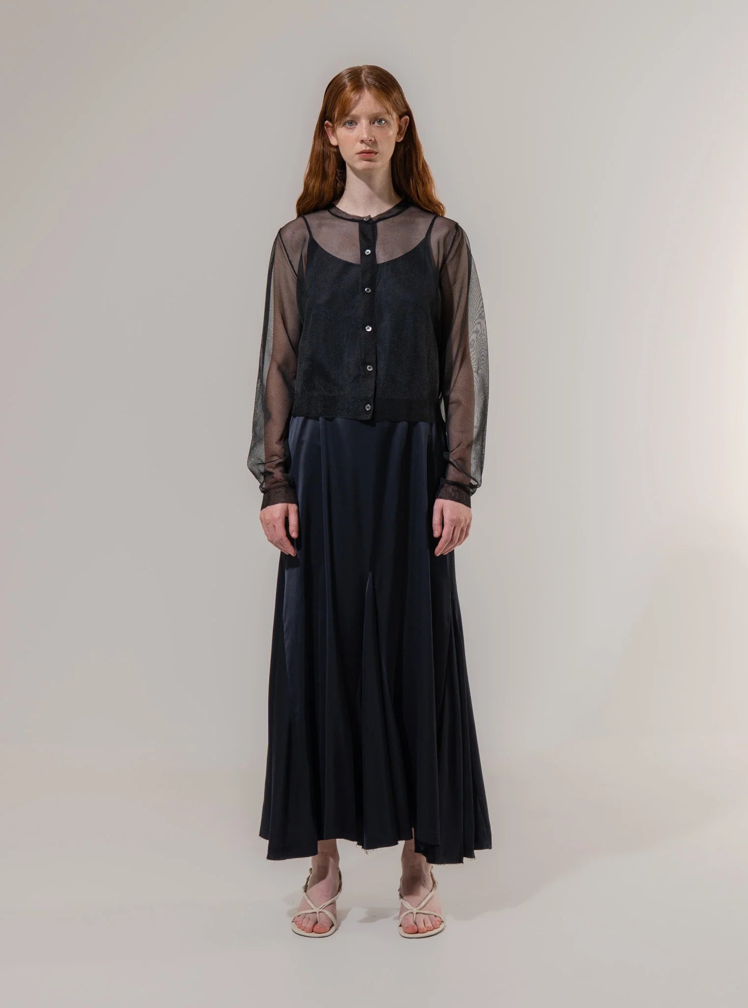 A woman stands facing forward wearing the Sheer Cardigan - Black by Emin and Paul over a black dress with white sandals, set against a plain light background.