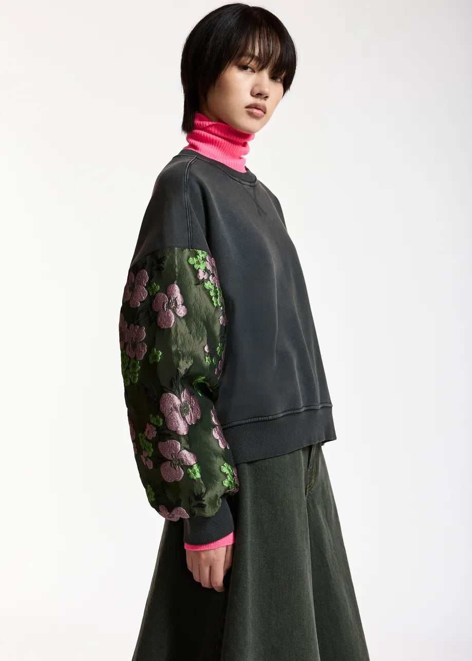 A model wearing a dark charcoal gray cropped sweatshirt with contrasting floral patterned sleeves, a bright pink turtleneck, and a dark green skirt, photographed against a plain white studio background.