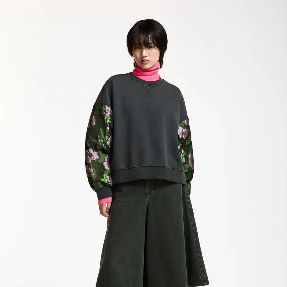 A model stands against a plain white studio background wearing a dark charcoal gray sweatshirt with green floral patterned sleeves, a bright pink turtleneck underneath, a dark olive green wide-leg skirt, black leggings, and black heeled boots.