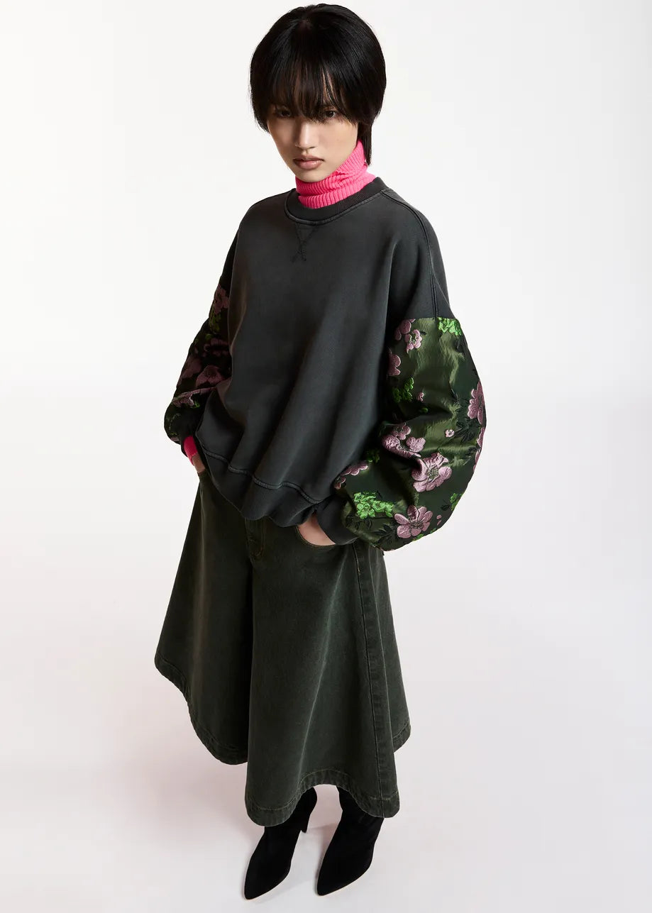 A model wearing a dark charcoal gray oversized sweatshirt with contrasting green floral patterned sleeves, paired with a dark green skirt and black pointed-toe heels, shot against a plain white studio background.