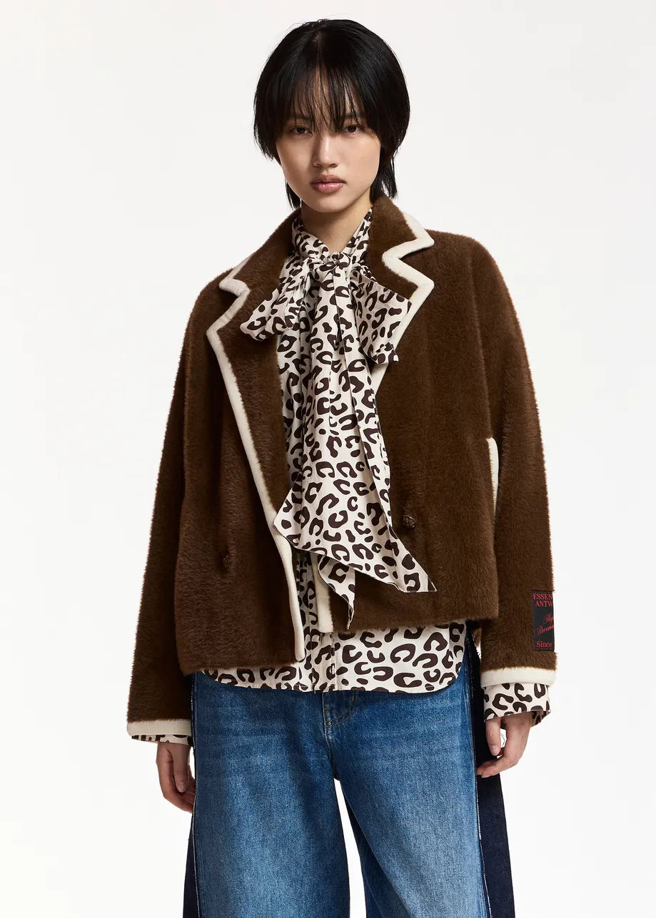 A model wearing a brown teddy-textured jacket with cream-colored piping, layered over a leopard-print blouse and blue jeans, against a plain white studio background.