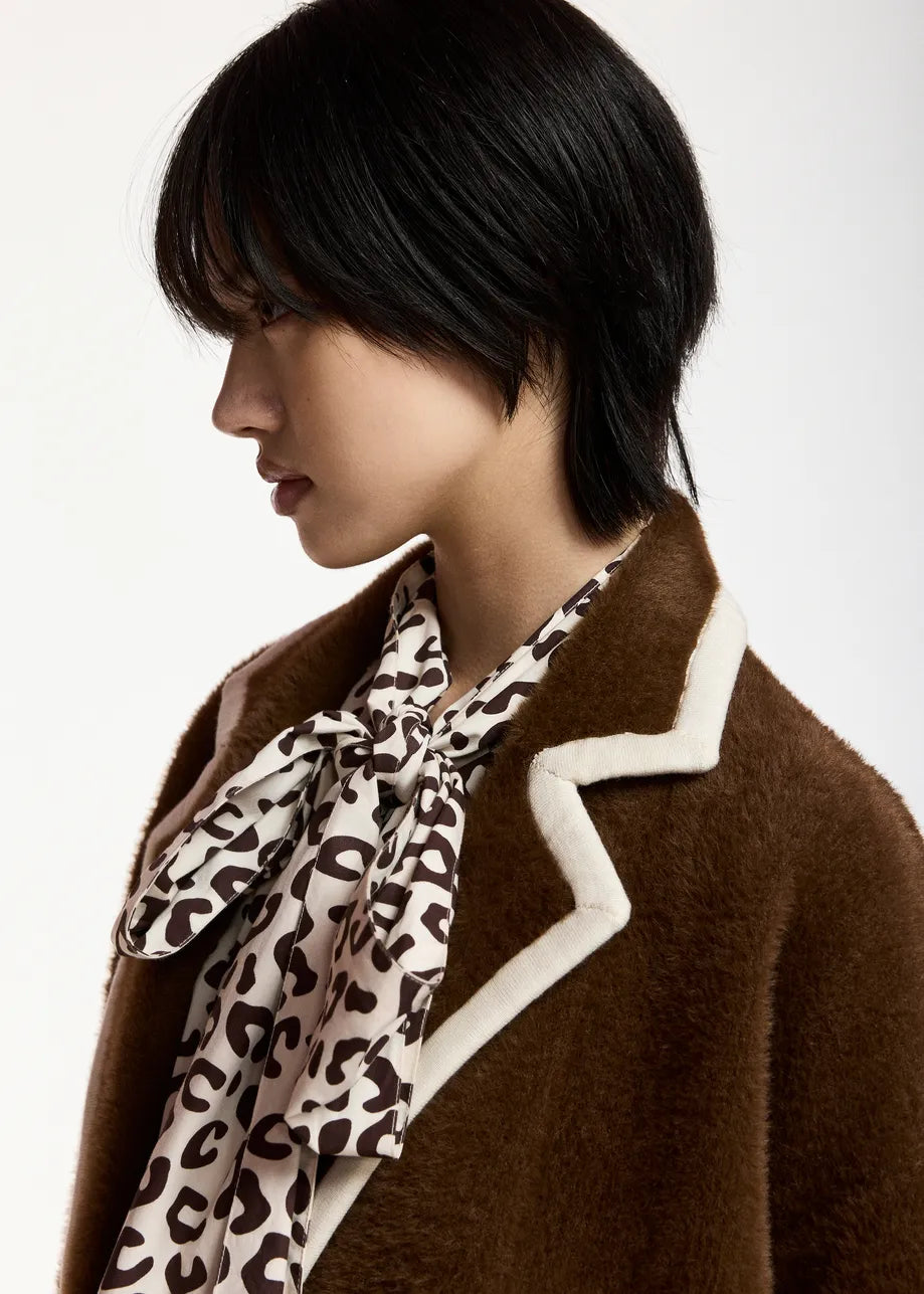 A close-up side profile of a model wearing a brown fuzzy coat with white contrast trim and a beige-and-brown leopard-print neck scarf, against a plain light background.
