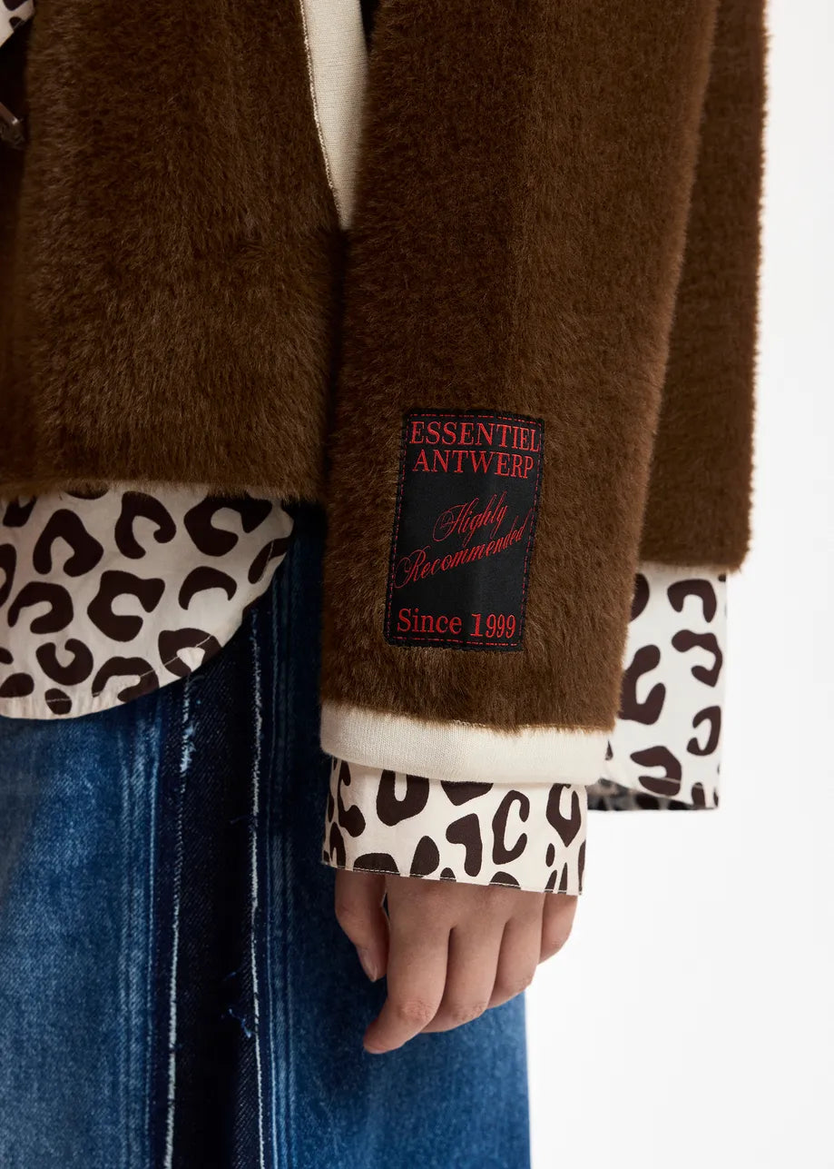 Close-up of a brown faux fur jacket with a cream lining and a black patch reading 'ESSENTIALS ANTWERP Style Statement Since 1989', worn over a leopard-print shirt and blue jeans.