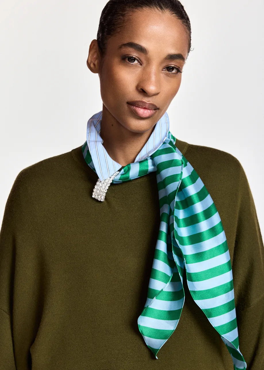 A model wearing a green and light blue striped silk scarf tied around a light blue striped shirt collar, paired with an olive green knit sweater, against a plain light gray background.
