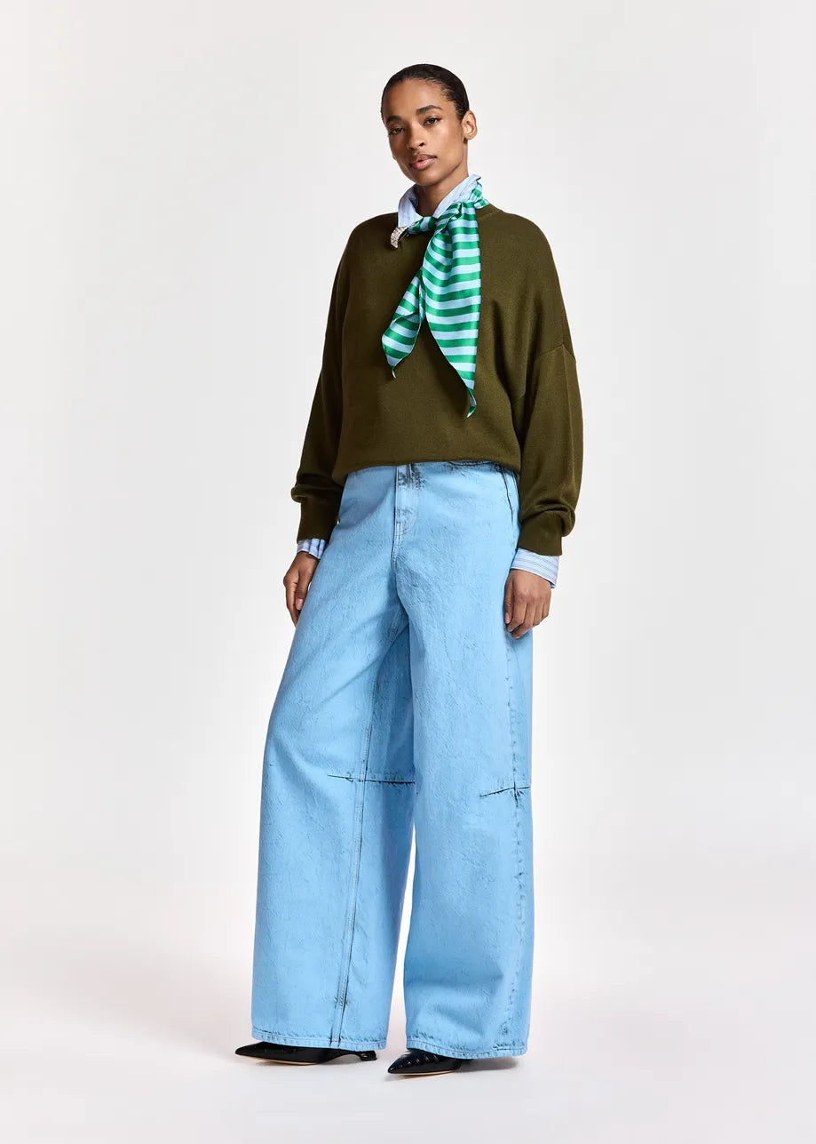 A model stands against a light gray studio background wearing a dark olive green long-sleeve top, light blue wide-leg jeans, a light blue collared shirt, a green and white striped neck scarf, and black shoes.