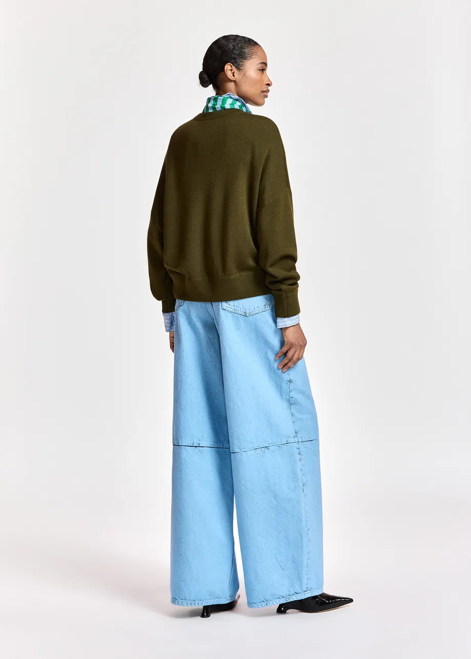 A person stands facing away, wearing the Essentiel Antwerp Kurtis Wool Blend Knit with Scarf over a green checked shirt and wide-leg light blue jeans, paired with black heeled shoes, against a plain light background.