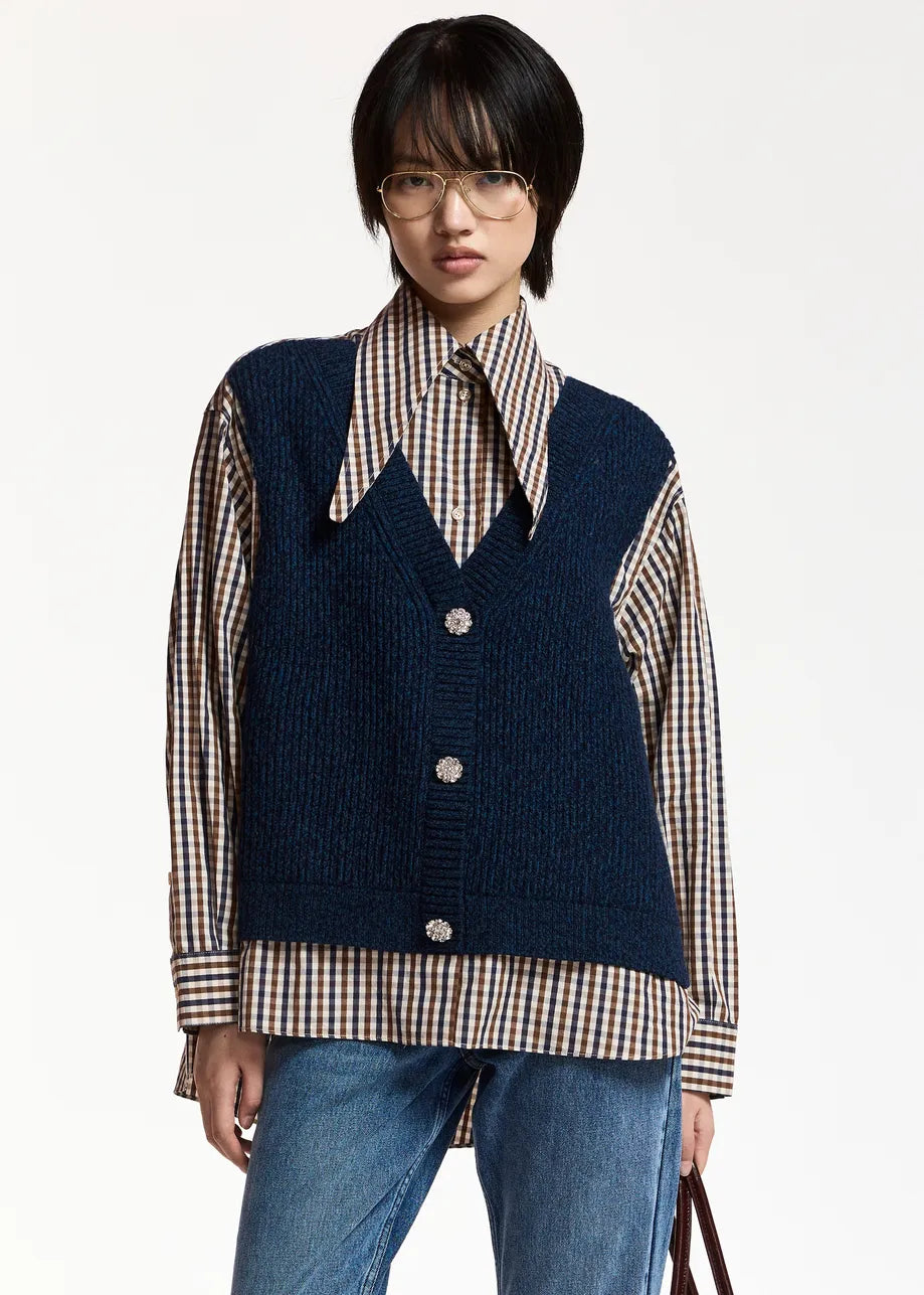 A person wearing glasses, a navy sweater vest layered over the Essentiel Antwerp Kute Fabric Mix Shirt—with its organic cotton checked design and wide collar—paired with blue jeans, standing against a plain white background.