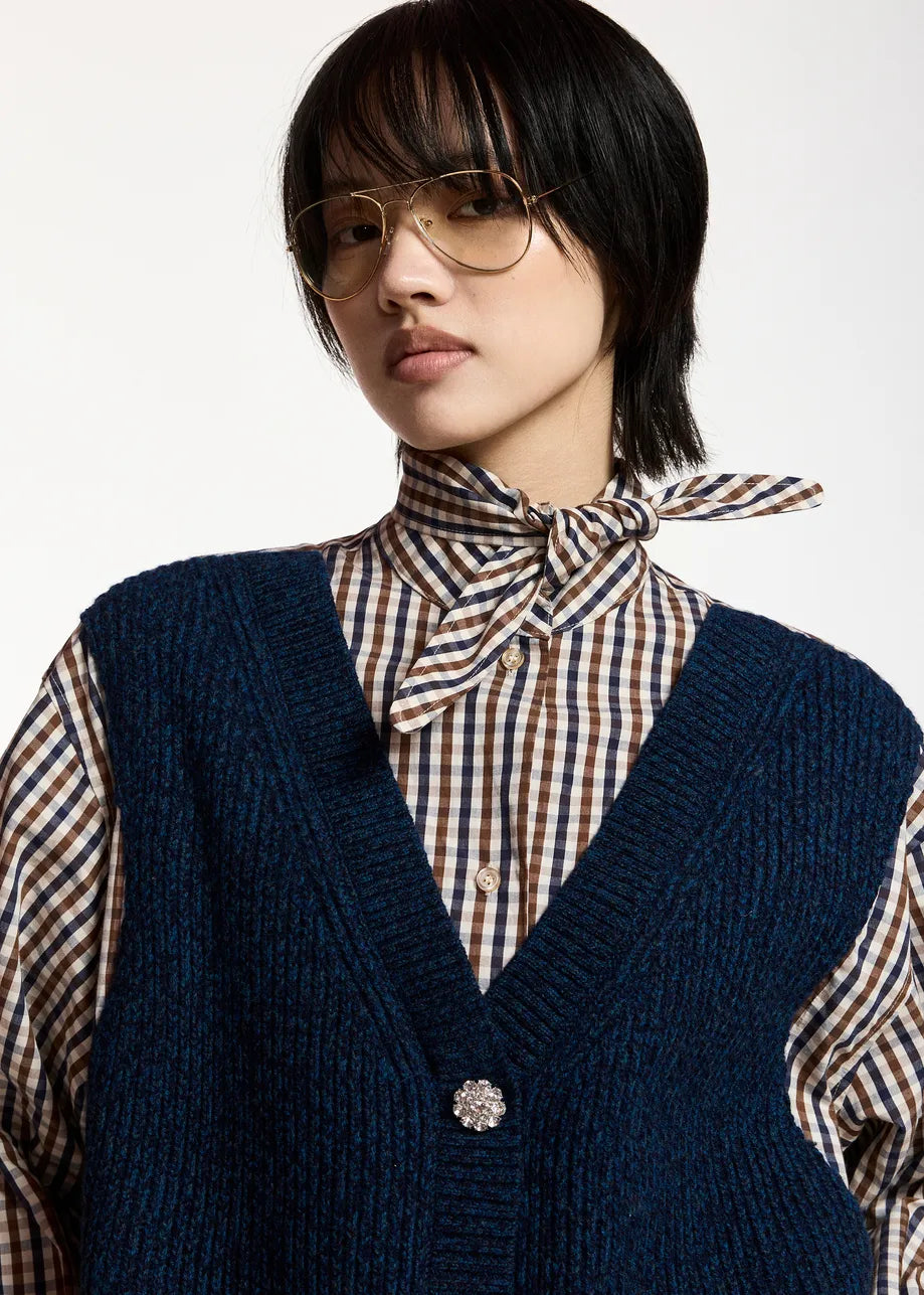 Person wearing glasses, the Essentiel Antwerp Kute Fabric Mix Shirt—an organic cotton checked button-up—styled with a necktie and a textured navy vest adorned with a brooch, looking at the camera against a plain background. The outfit features a subtly deconstructed layered effect, creating an effortlessly oversized silhouette.