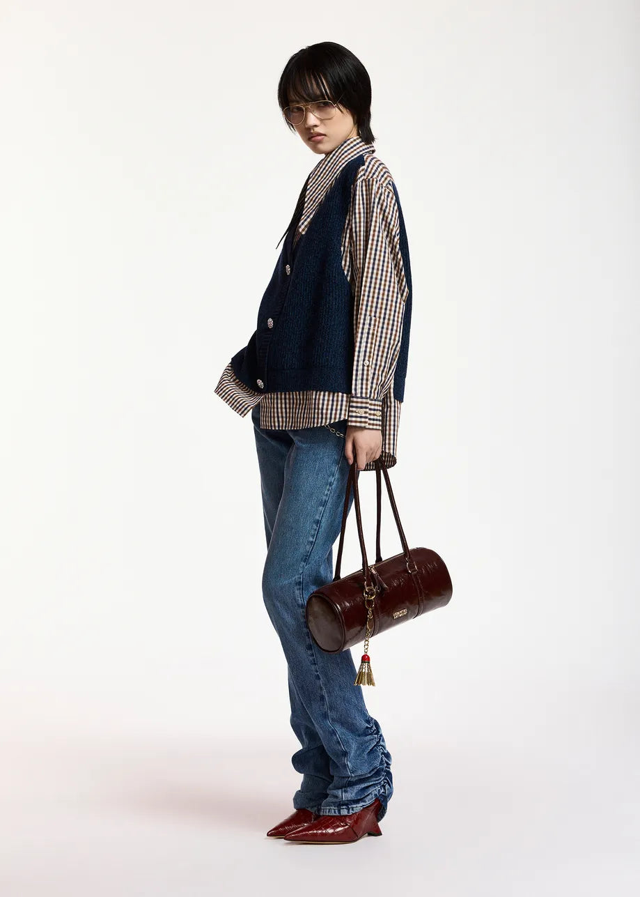 A person stands in profile against a plain white background, wearing the Essentiel Antwerp Kute Fabric Mix Shirt—an organic cotton checked piece—styled with a navy vest, blue jeans, and red pointed shoes. The look features an oversized silhouette and is completed with a brown cylindrical handbag, adding modern flair to the classic combination.