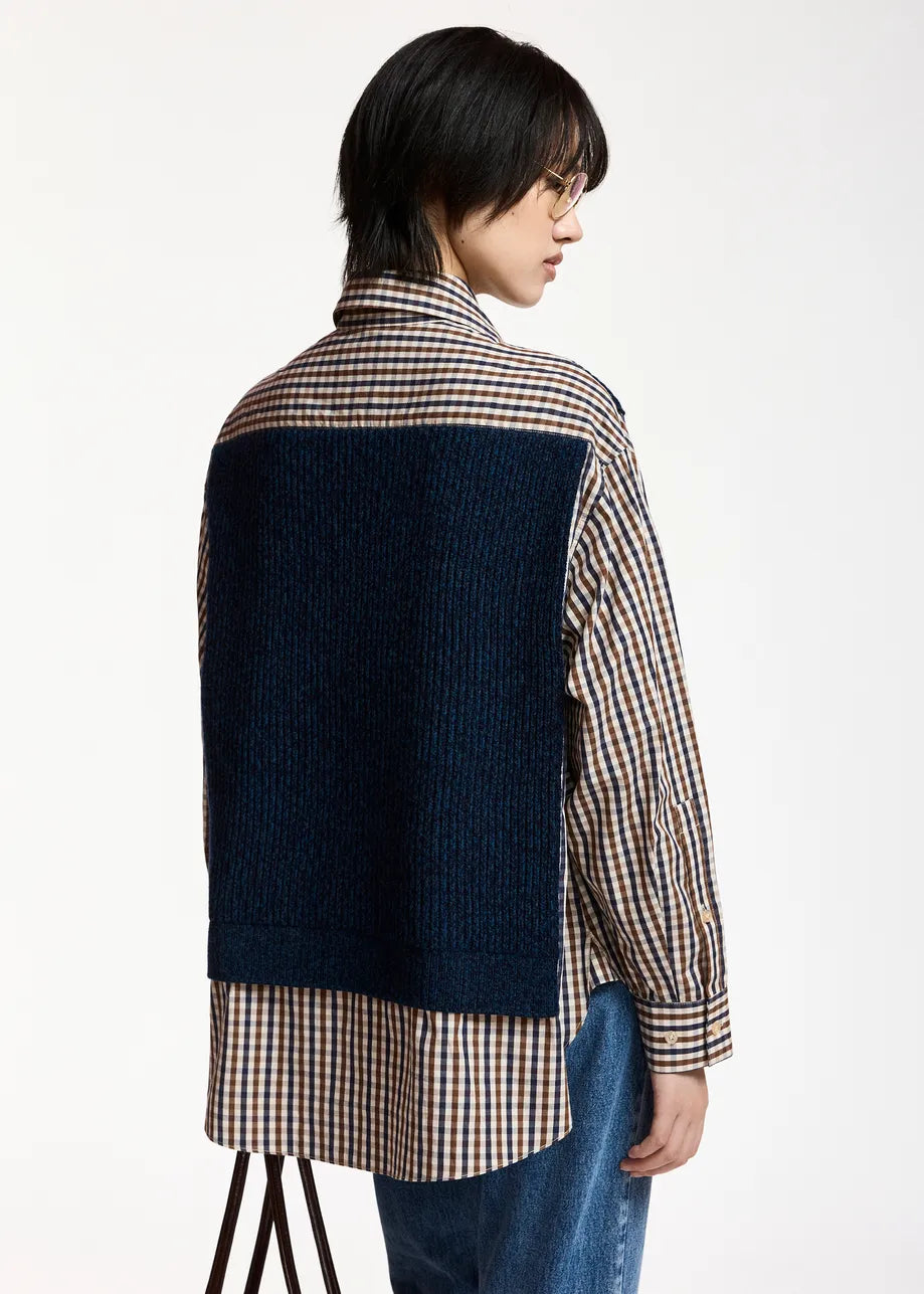 A person stands sideways against a plain white background, wearing the Essentiel Antwerp Kute Fabric Mix Shirt—an organic cotton checked piece—layered with a dark blue textured vest for a deconstructed effect, paired with blue jeans.