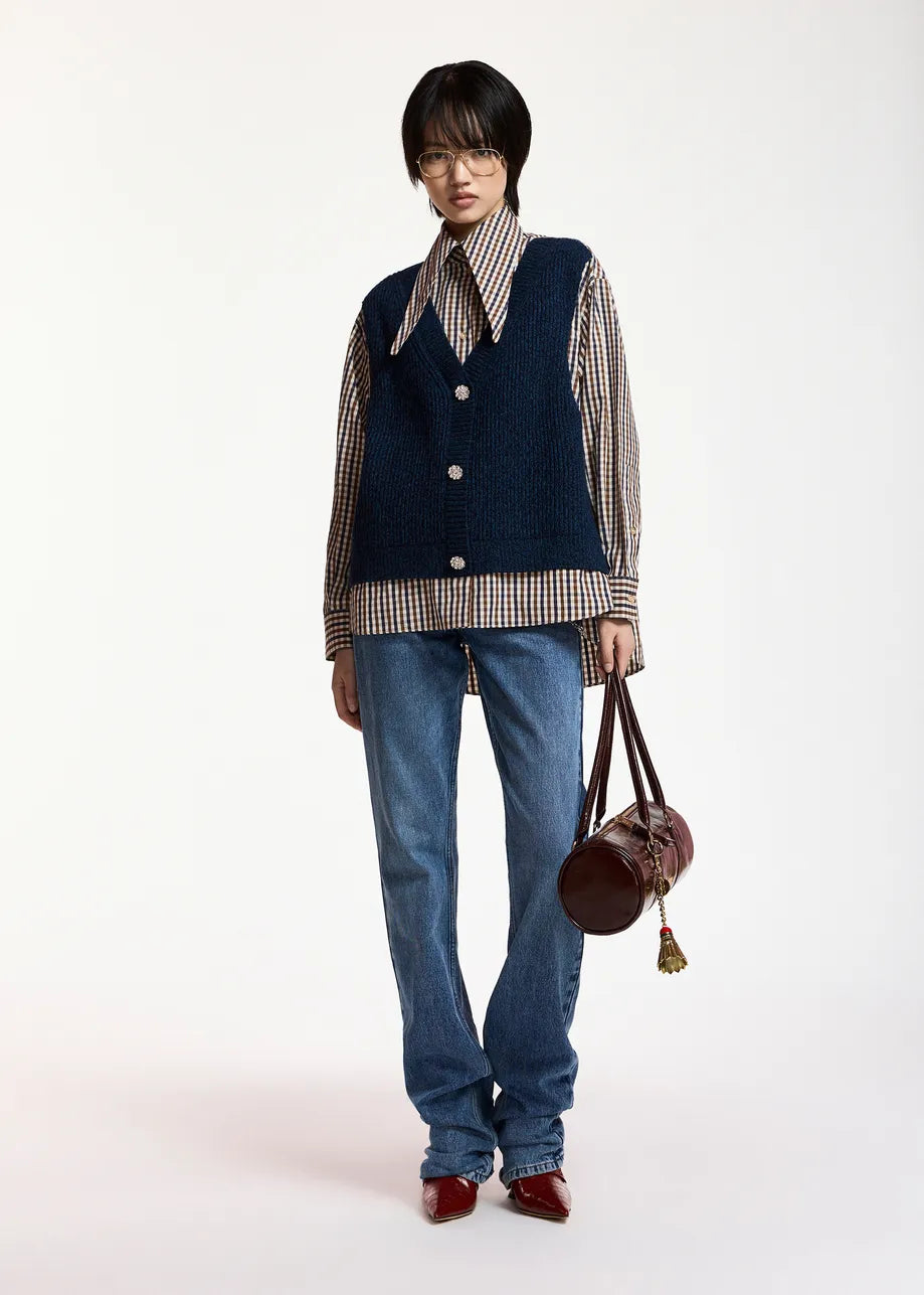 Wearing the Essentiel Antwerp Kute Fabric Mix Shirt, a person stands against a plain white background, styled with a navy vest, blue jeans, red shoes, and holding a brown round handbag.