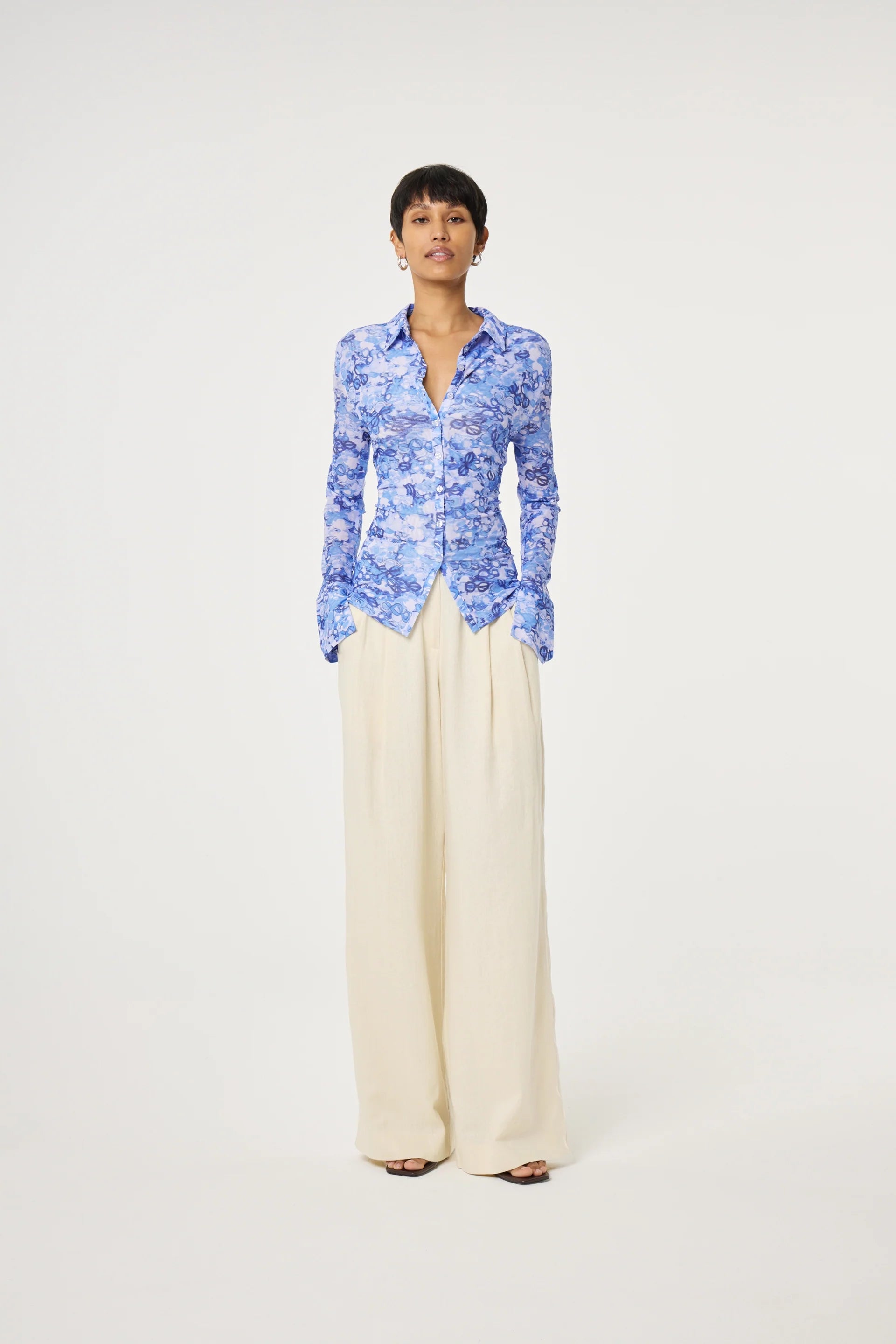 A person stands against a white background, wearing the Fabienne Chapot Kevis Blouse - Blue, a bright blue floral button-up with a fitted silhouette, paired with wide-legged cream pants and hands in their pockets.