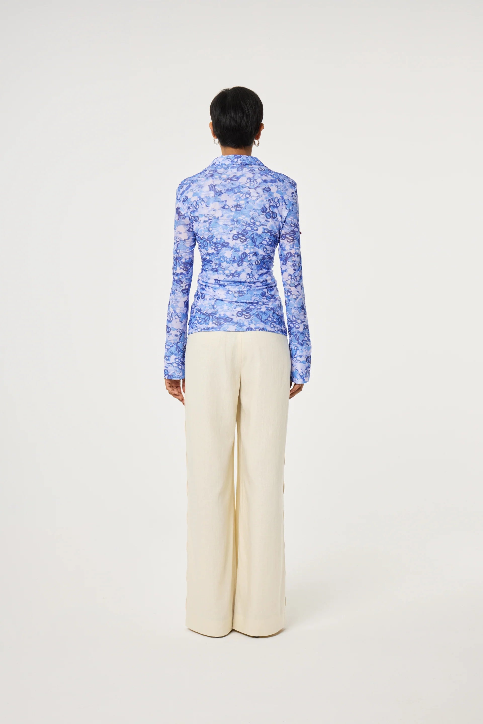 Person standing with their back to the camera, wearing a Fabienne Chapot Kevis Blouse - Blue with a fitted silhouette and wide-leg cream pants against a plain white background.
