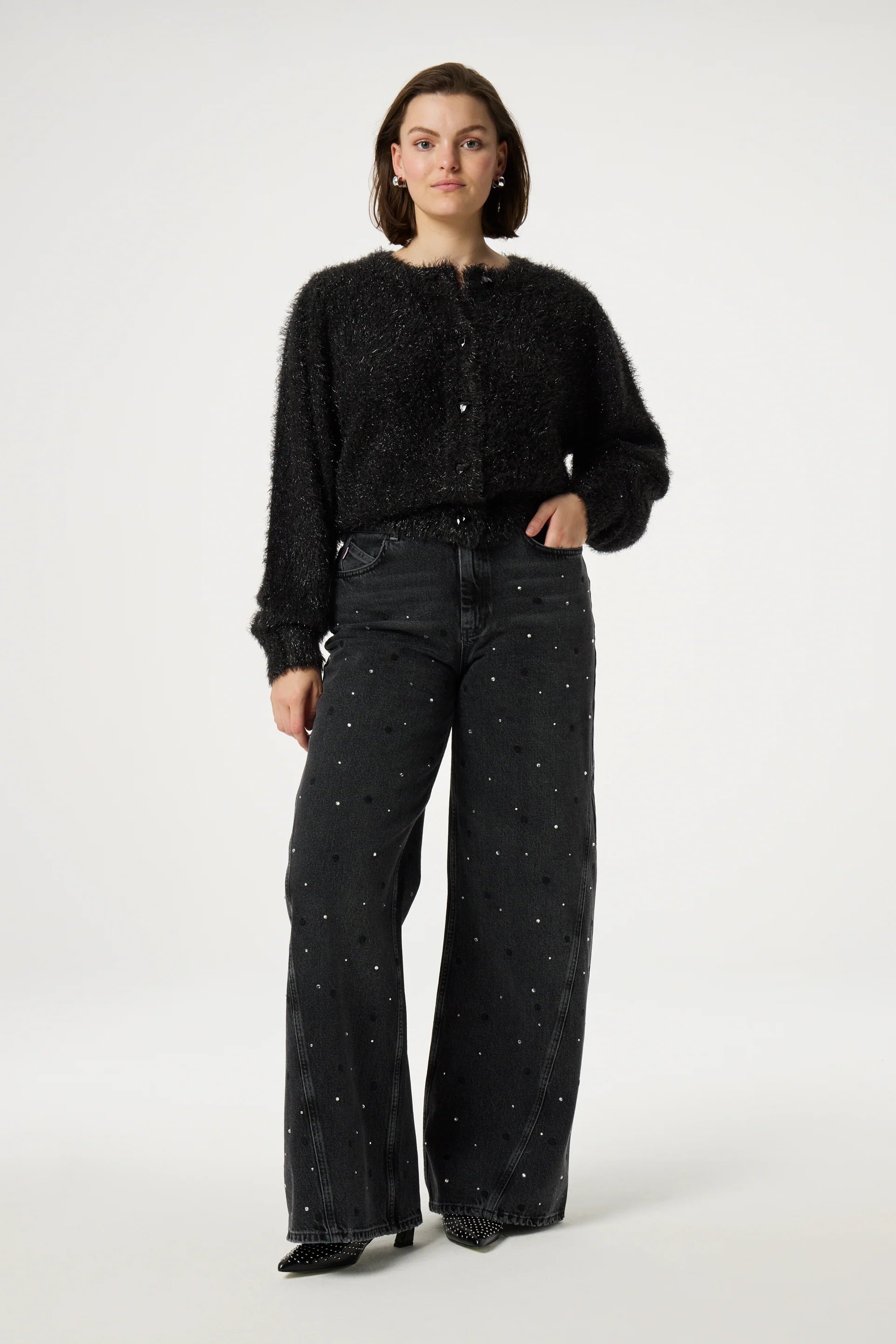 A person stands facing forward, embracing the sparkle season in the chic Kitty Cardigan - Black by Fabienne Chapot, paired with wide-leg jeans adorned with subtle rhinestone embellishments.