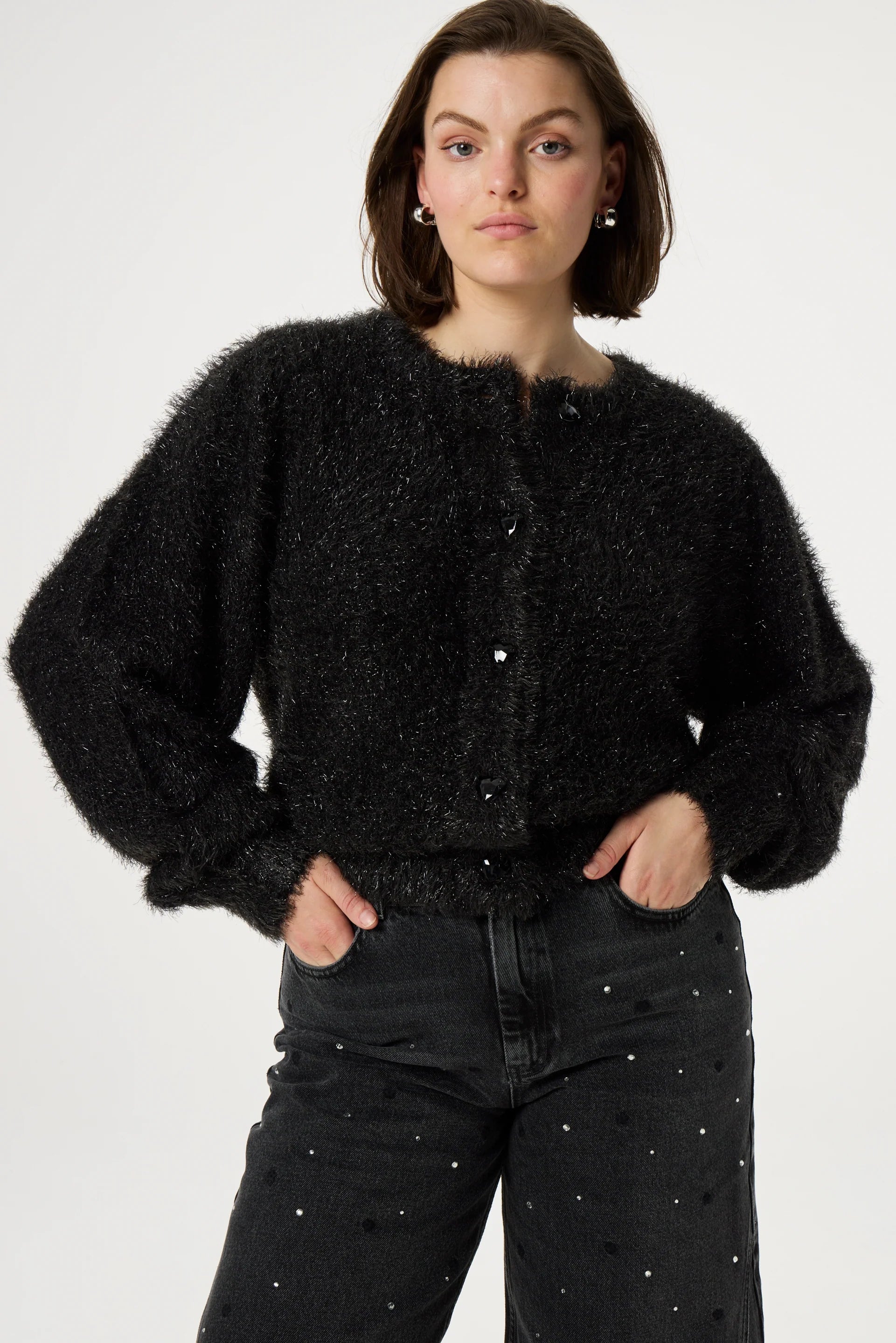 A person wearing the Kitty Cardigan in black by Fabienne Chapot, featuring heart-shaped buttons, and dark pants with small embellishments stands against a plain background.