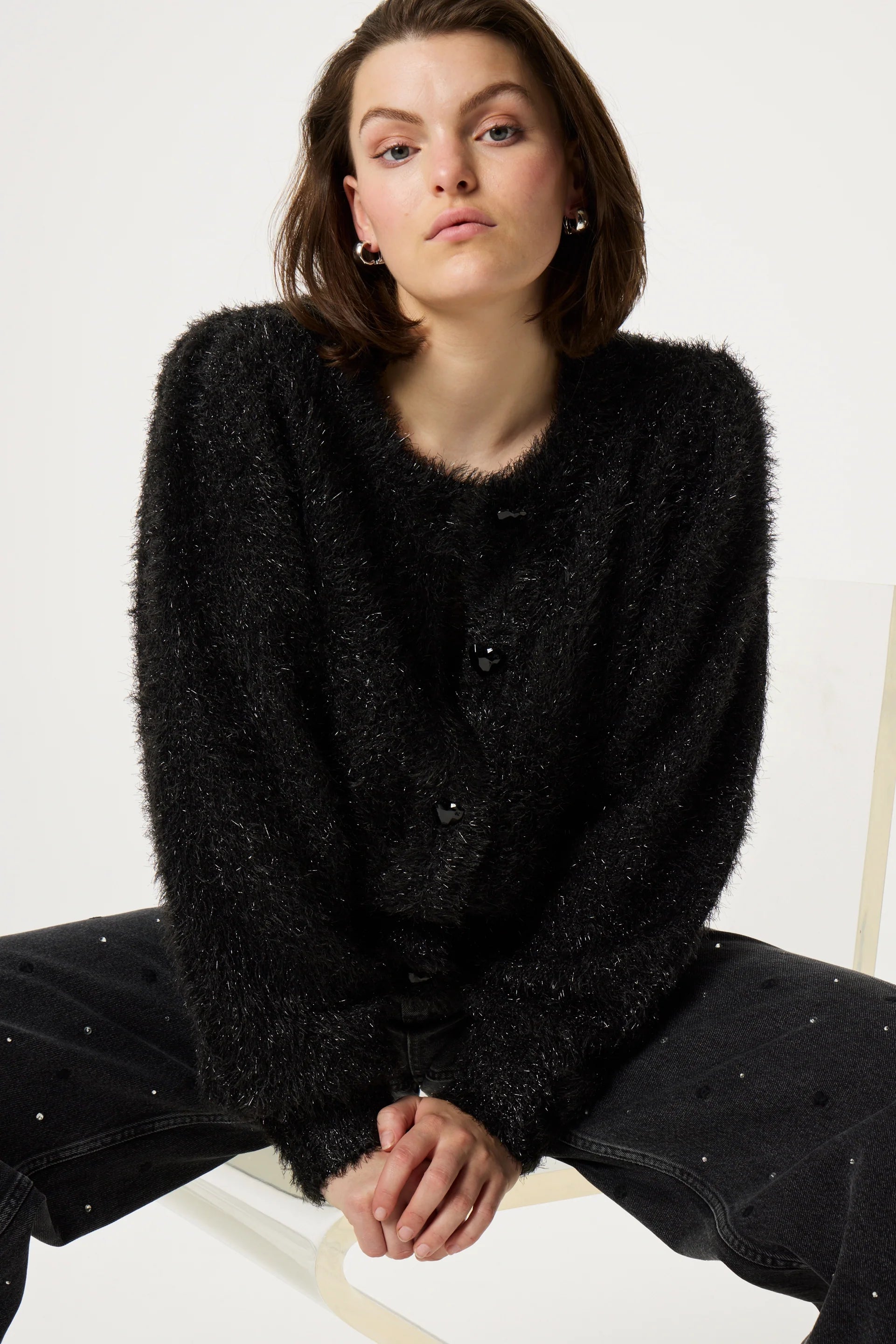 A person with short brown hair is sitting in a relaxed posture, wearing the Fabienne Chapot Kitty Cardigan in black, featuring heart-shaped buttons, paired with dark jeans.