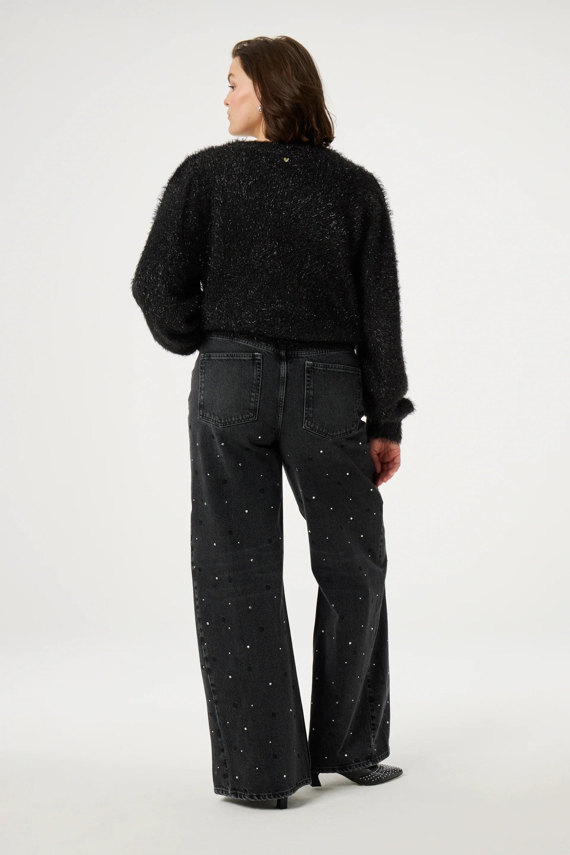 A person wearing the Kitty Cardigan - Black by Fabienne Chapot, featuring heart-shaped buttons, pairs it with dark wide-leg pants accented with small embellishments while standing against a plain background and facing away.