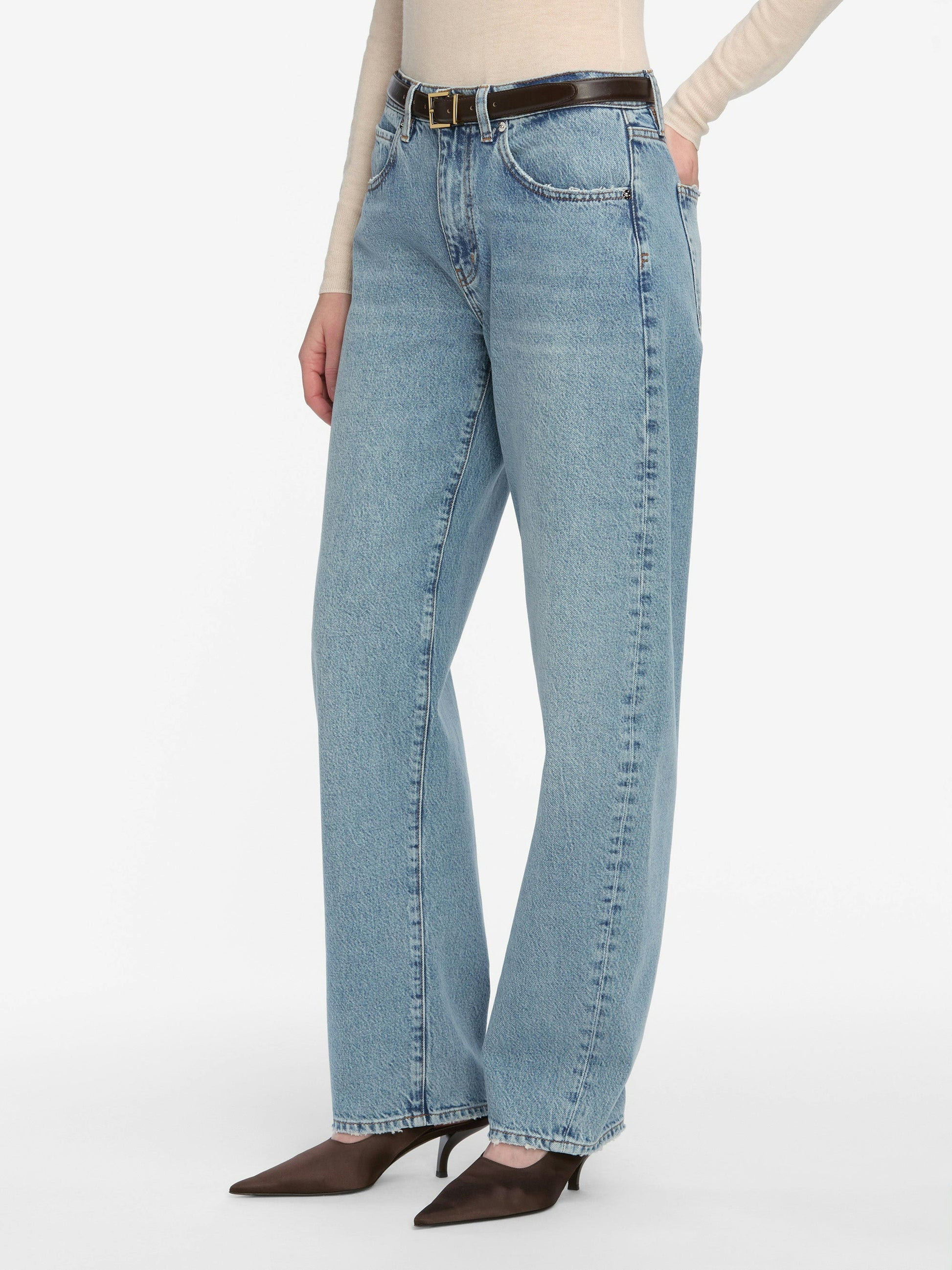 A person wears Frame's Low Slung Barrel - Dollhouse light blue jeans with a beige long-sleeve top, black belt, and pointed brown shoes, standing hands-in-pockets against a plain background.
