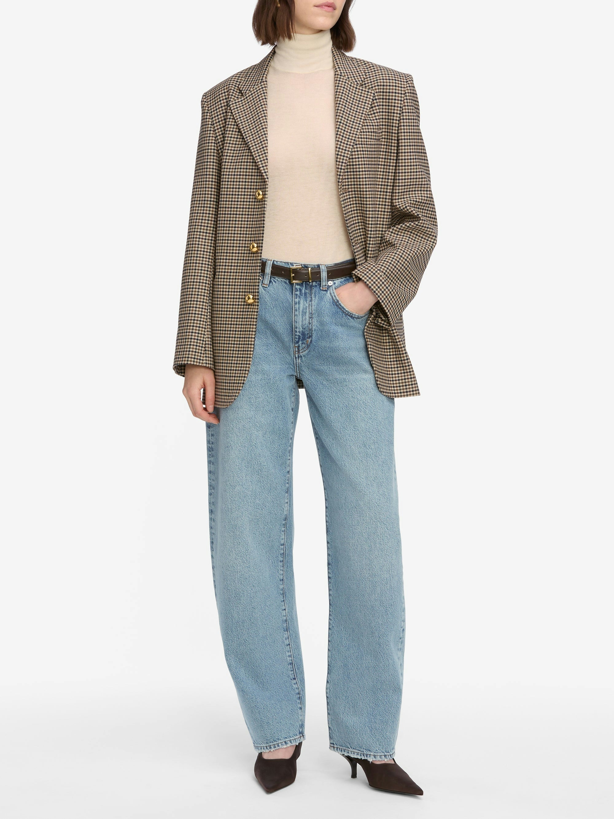 Person wearing Frame’s Low Slung Barrel - Dollhouse jeans, a beige turtleneck, checked blazer, black belt, and dark pointed-toe heels, posing against a plain white background.