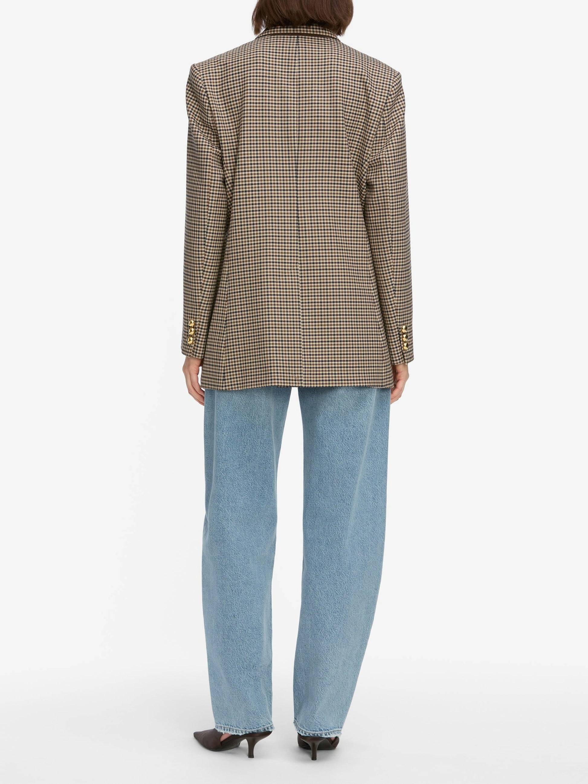 Person stands with back to camera, wearing the Frame Low Slung Barrel - Dollhouse jeans, a checkered blazer, and dark heeled shoes against a plain white background.