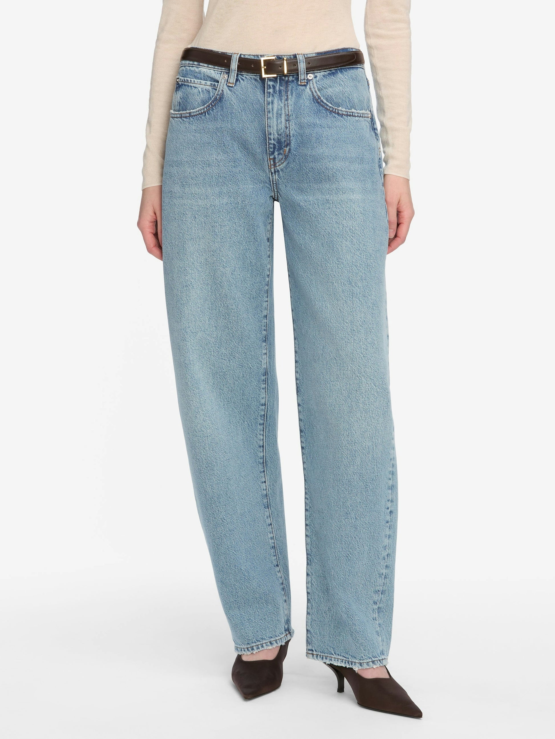 Model in FRAME’s Low Slung Barrel - Dollhouse jeans styled with a black belt, beige long-sleeve top, and dark pointed-toe heels, showcasing a trending silhouette against a white background.