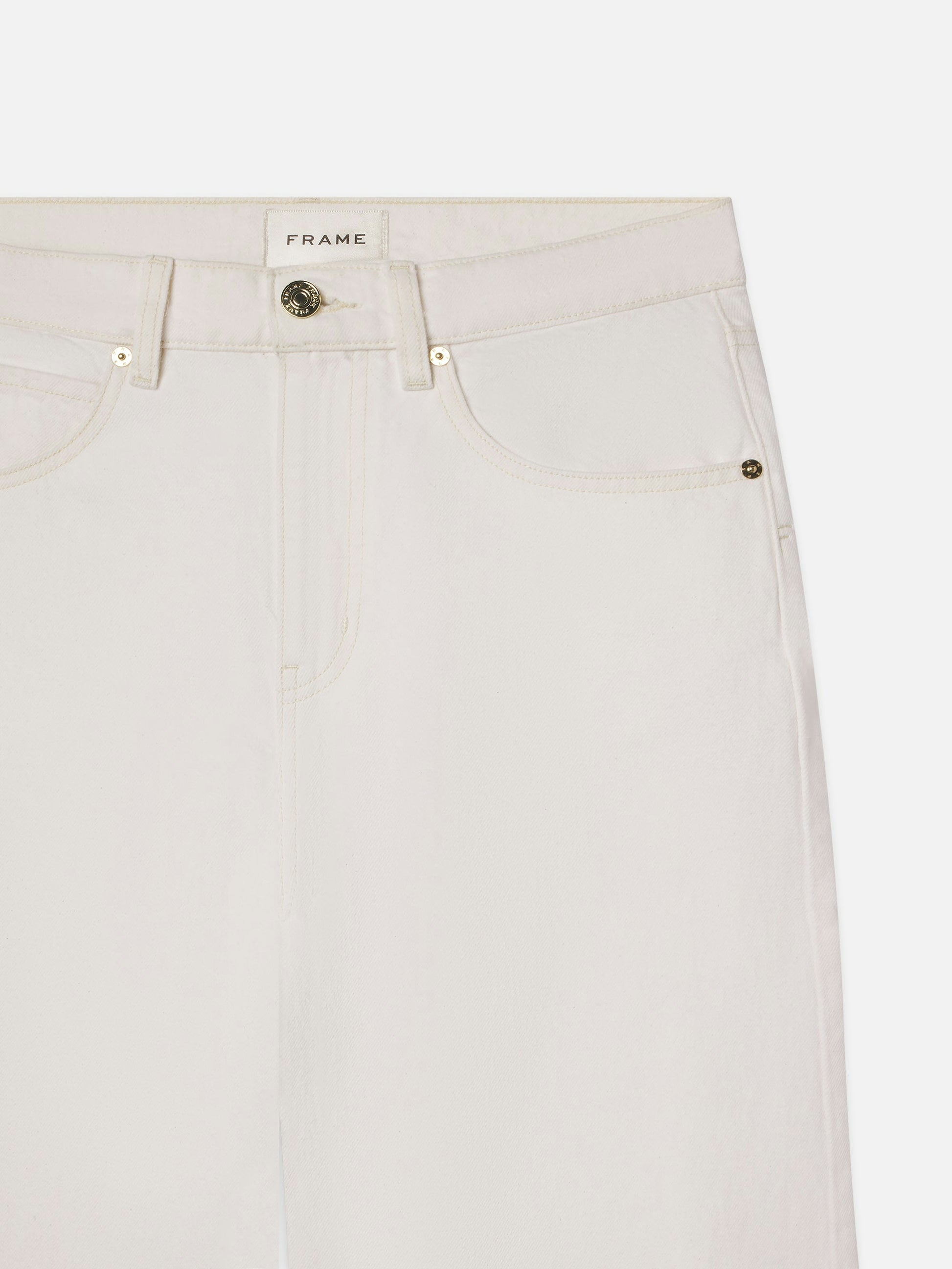 A close-up of the "Low Barrel Leg - Au Natural Clean" jeans by Frame shows 100% cotton, high-waisted design in off-white with a button fly and front pockets, featuring a branded label at the waistband.