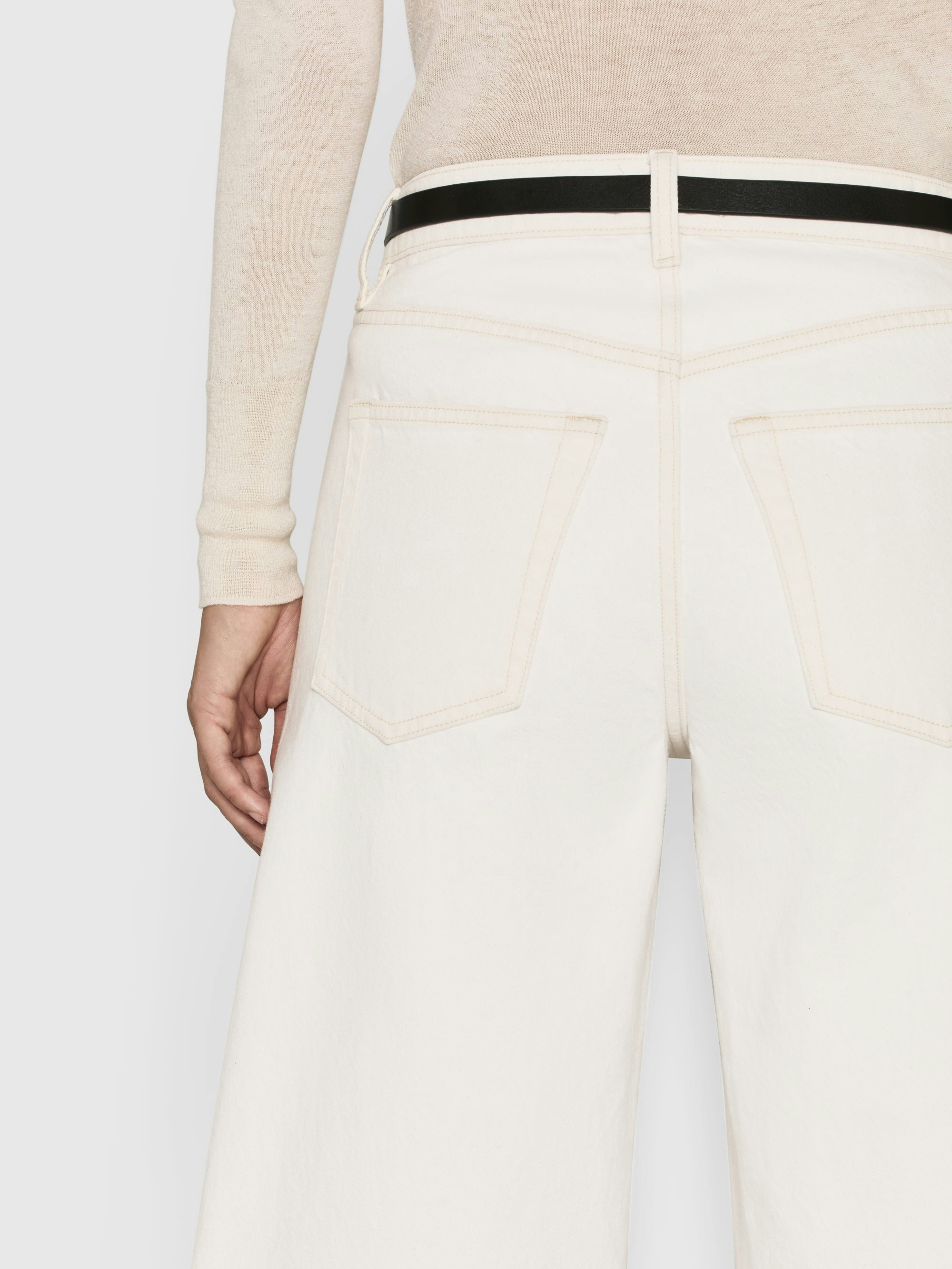 Rear view of a person wearing Frame's Low Barrel Leg - Au Natural Clean, featuring back pocket details and a black belt, highlighting the beige sweater paired with white wide-leg jeans made from 100% cotton, low-rise fit.