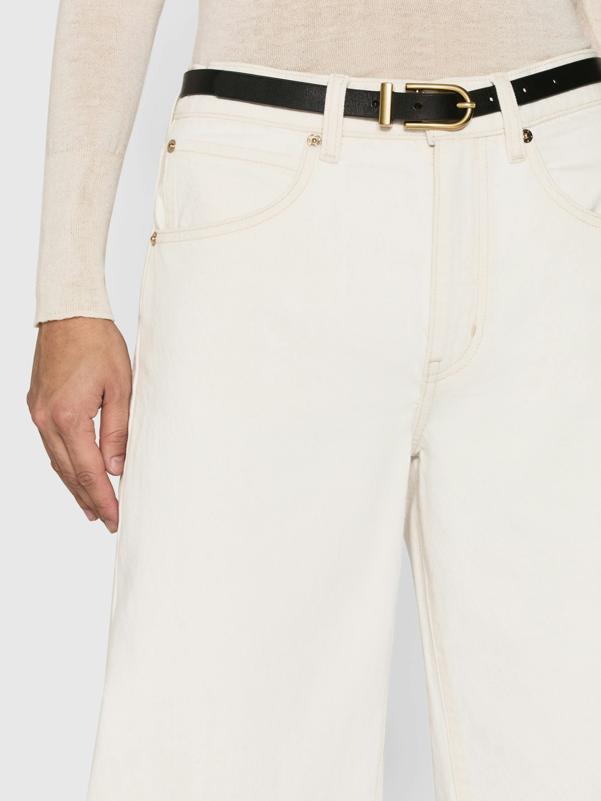 Dressed in Frame's Low Barrel Leg - Au Natural Clean 100% cotton jeans with a low rise, accessorized with a black belt, the person casually rests their left hand near the pocket against a plain background.