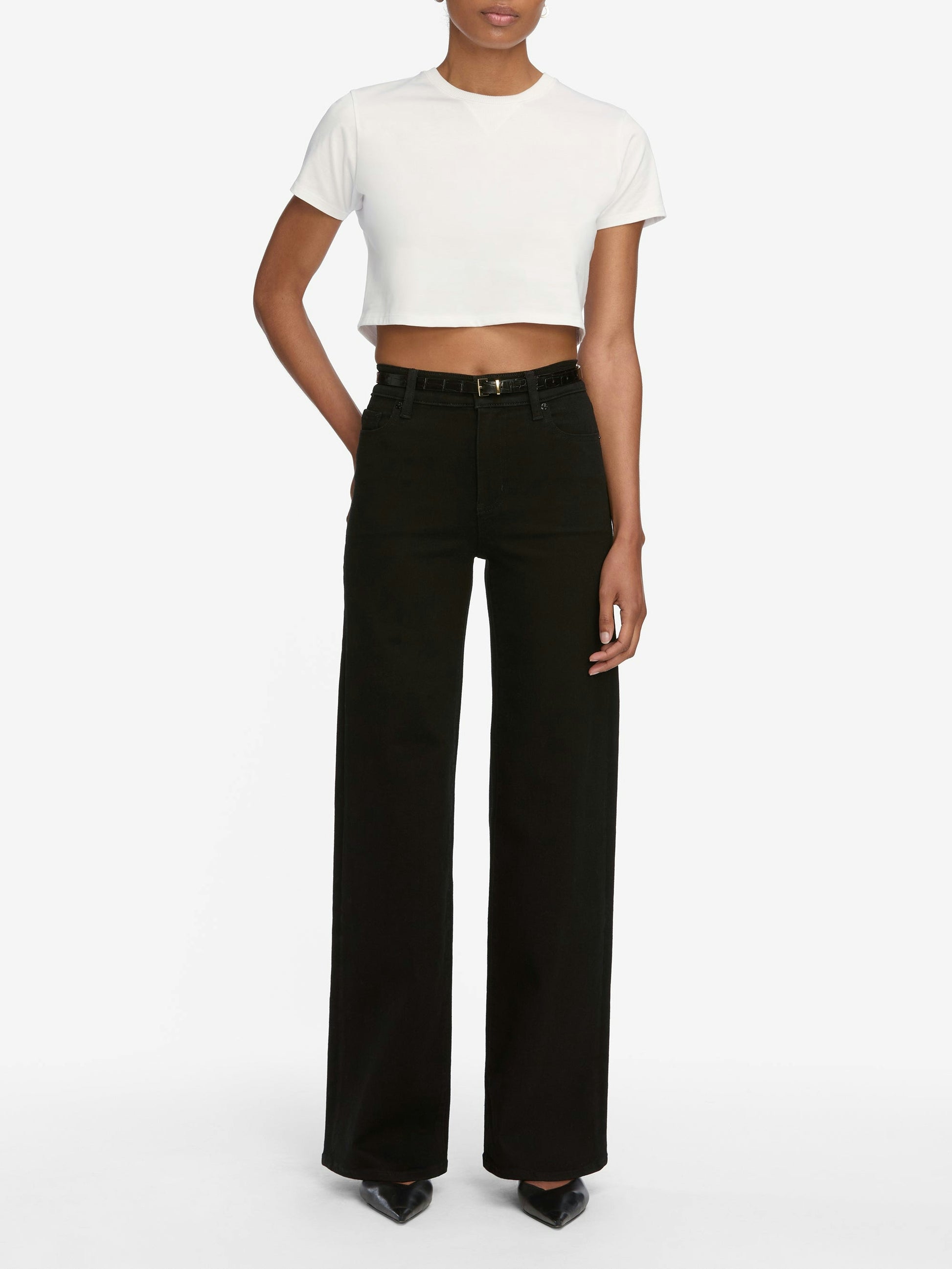 A person wearing a white short-sleeve crop top, Frame Le Slim Palazzo Jeans in black by Frame, and black pointed shoes stands against a plain white background.