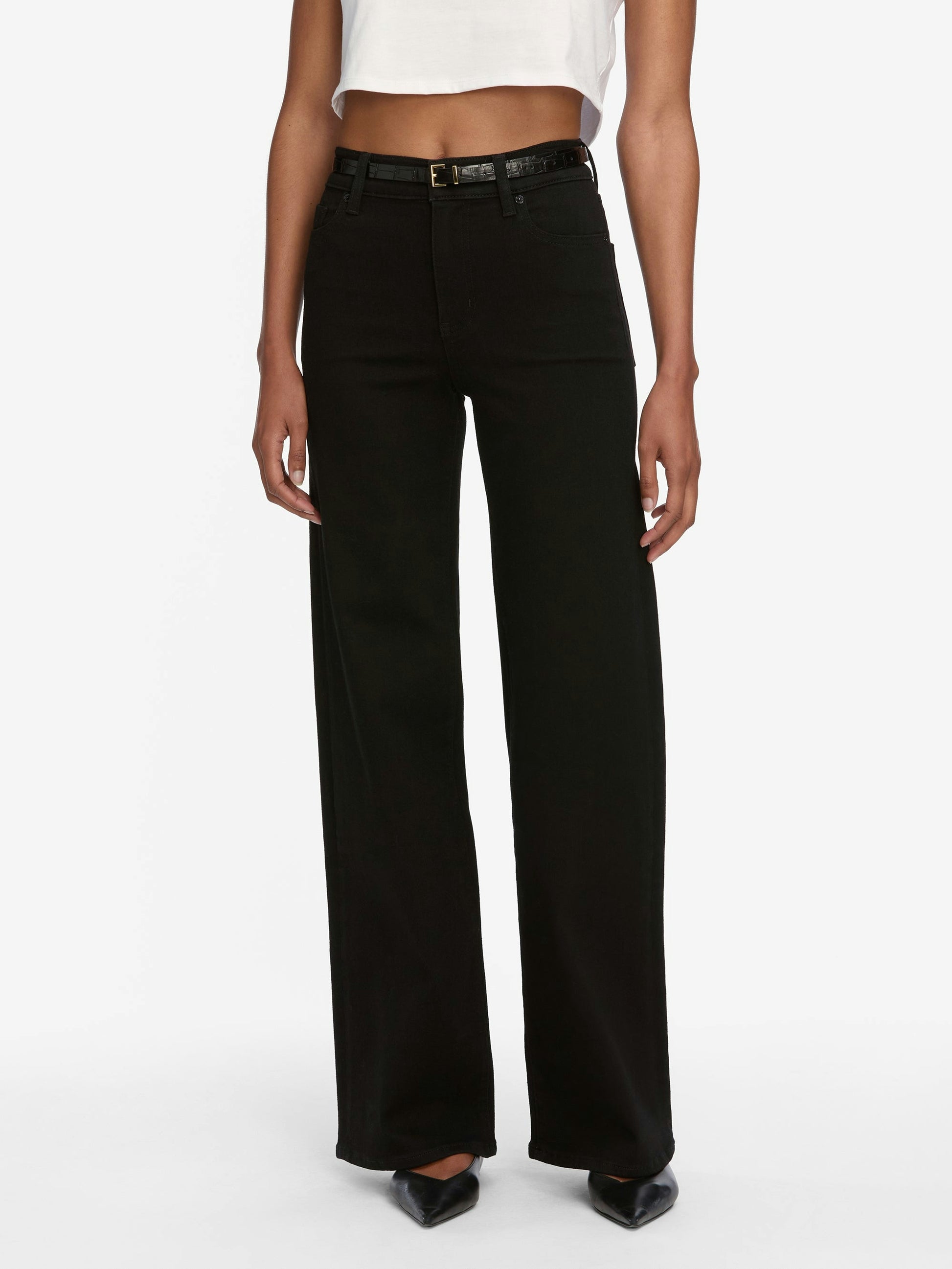 A person wearing Frame Le Slim Palazzo Jeans in black by Frame, styled with a black belt, white crop top, and black pointed shoes, stands against a plain background.