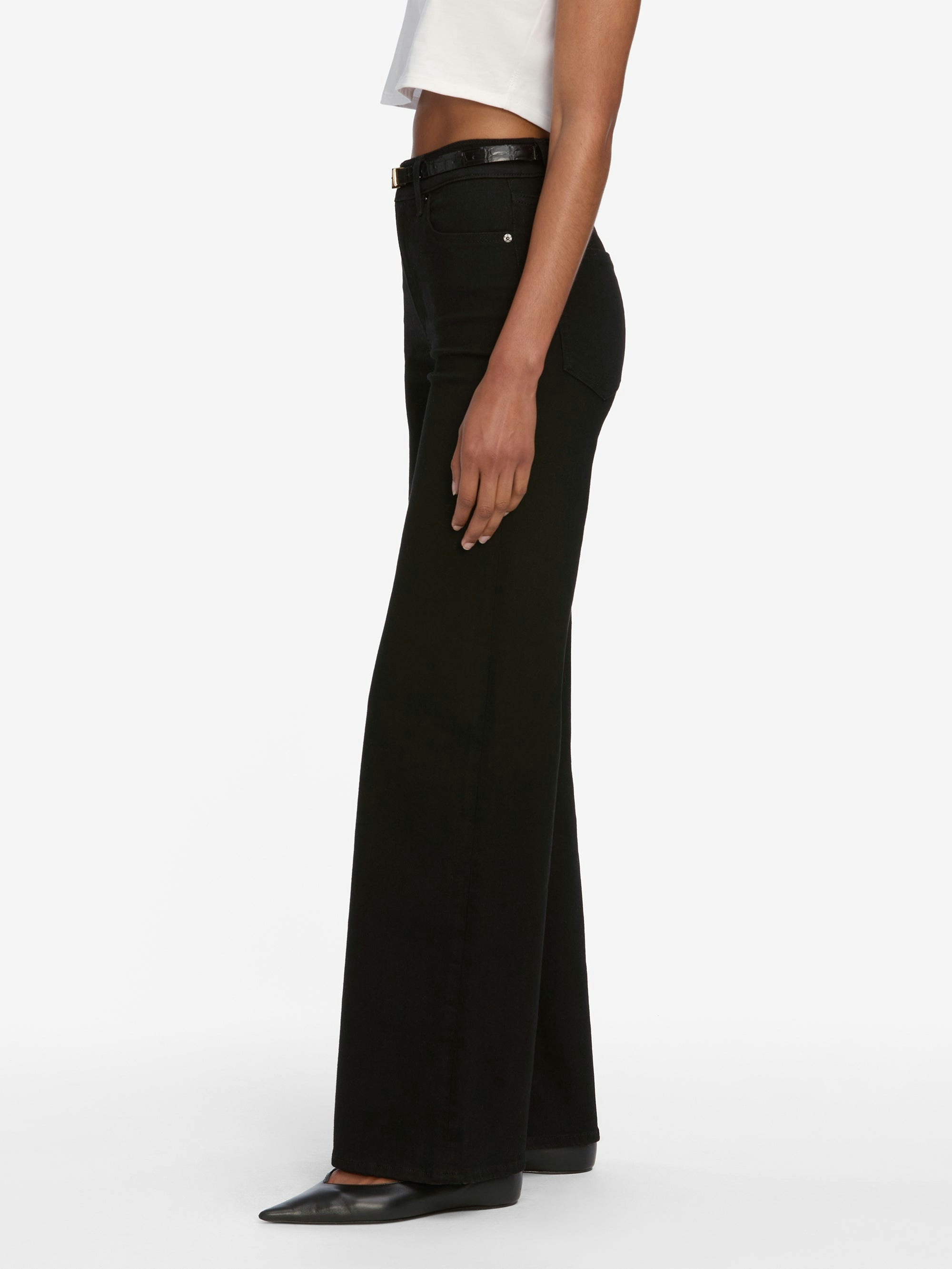 A person wearing Frame Le Slim Palazzo Jeans - Black by Frame, styled with a white cropped top and black pointed shoes, stands against a plain white background.